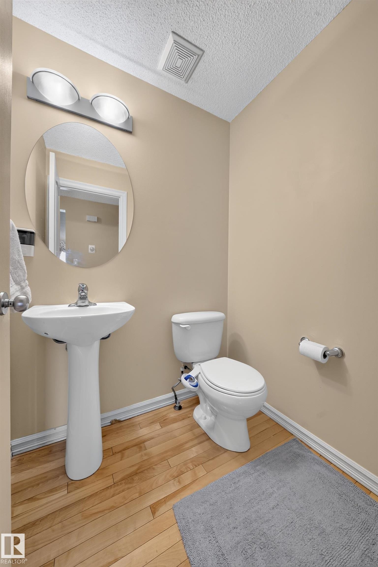 Pedestal sink and toilet with light wood-finish flooring, a round wall mirror, and a two-light vanity fixture - 2518 33A Avenue, Edmonton, AB