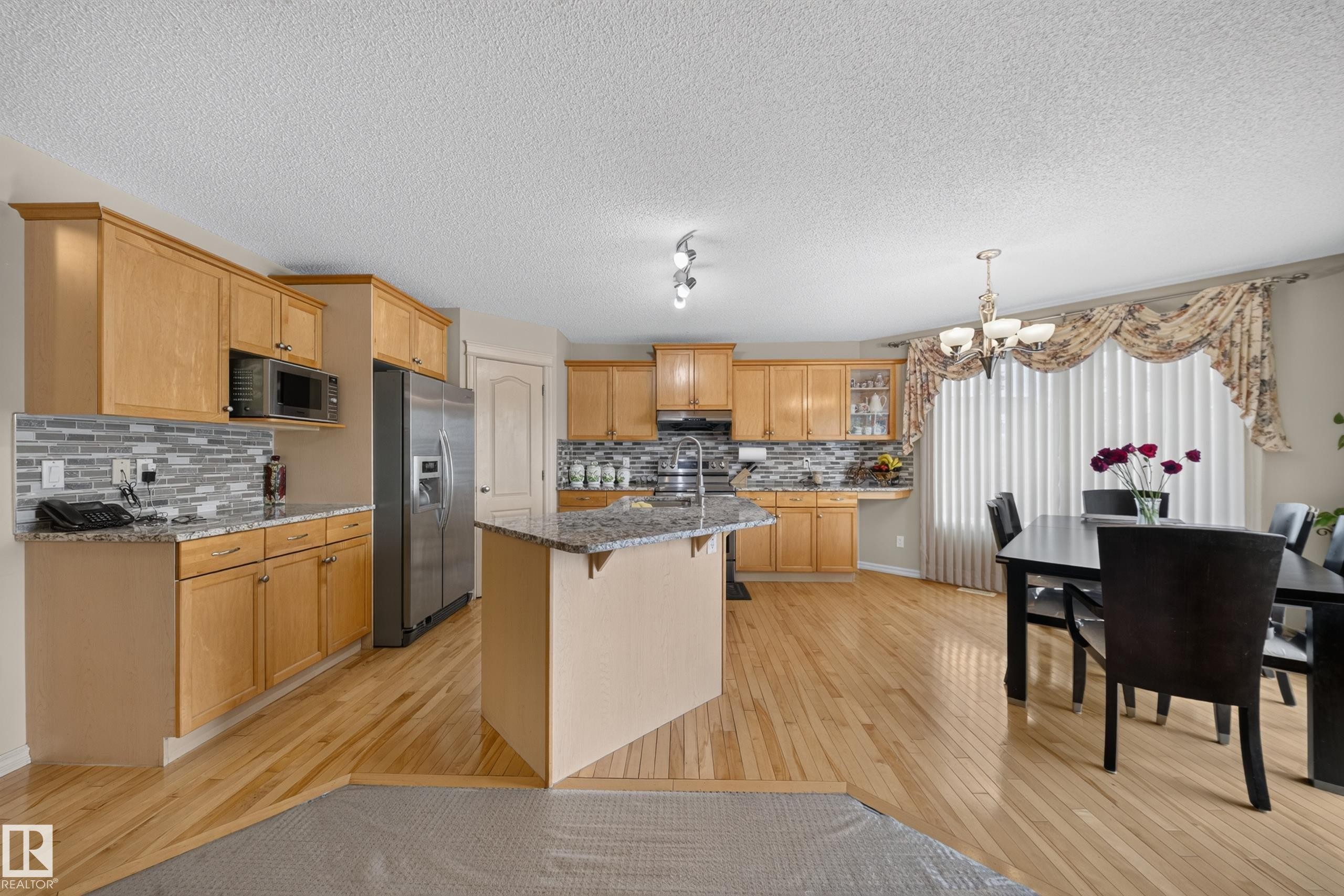 Spacious kitchen and dining area featuring wood-finish flooring, light-toned cabinetry, stainless steel appliances, a center island with a granite countertop, and a tiled backsplash - 2518 33A Avenue, Edmonton, AB