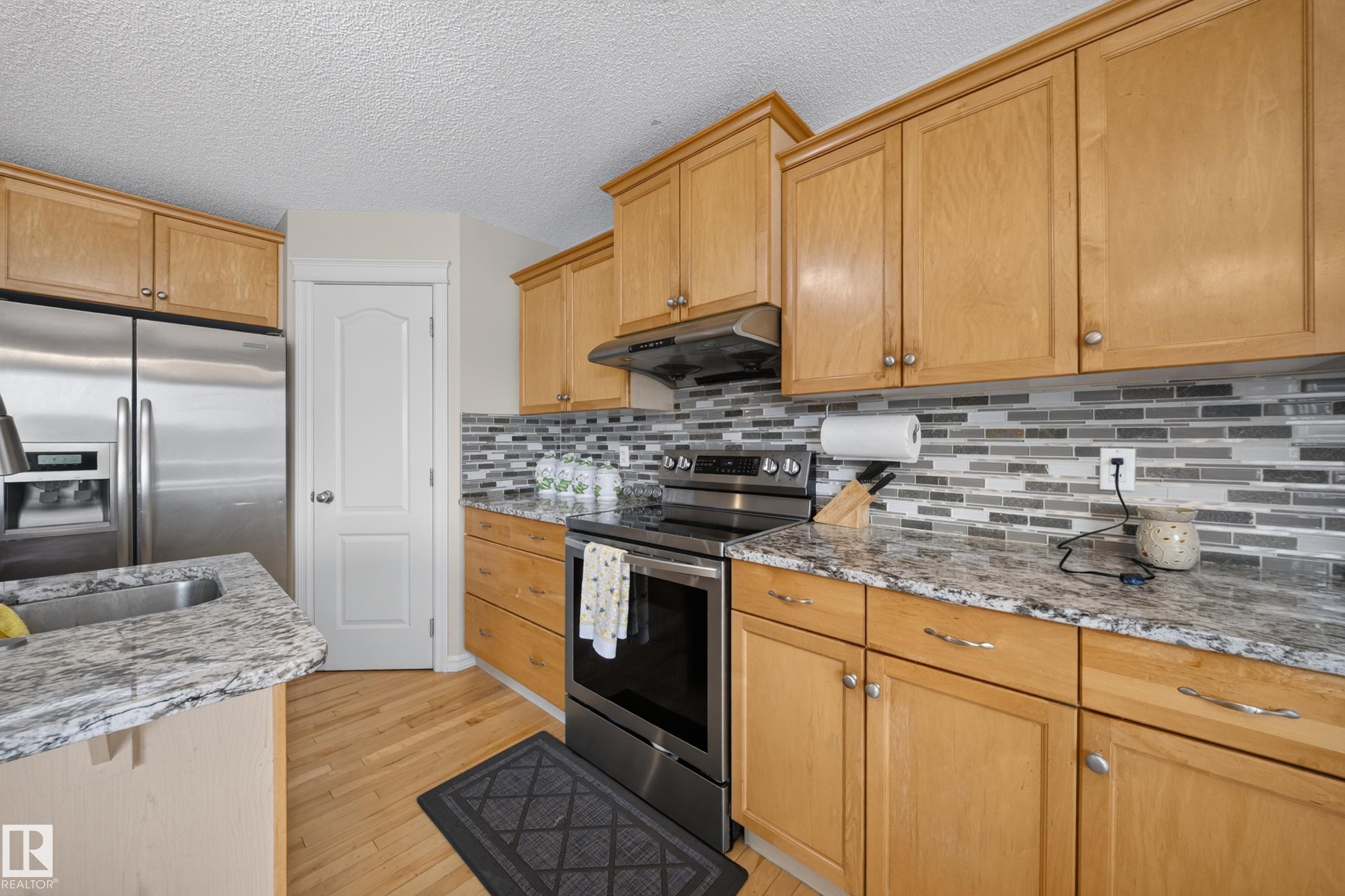 Kitchen featuring wood cabinetry, stainless steel appliances, speckled countertops, a mosaic tile backsplash, and wood-finish flooring - 2518 33A Avenue, Edmonton, AB