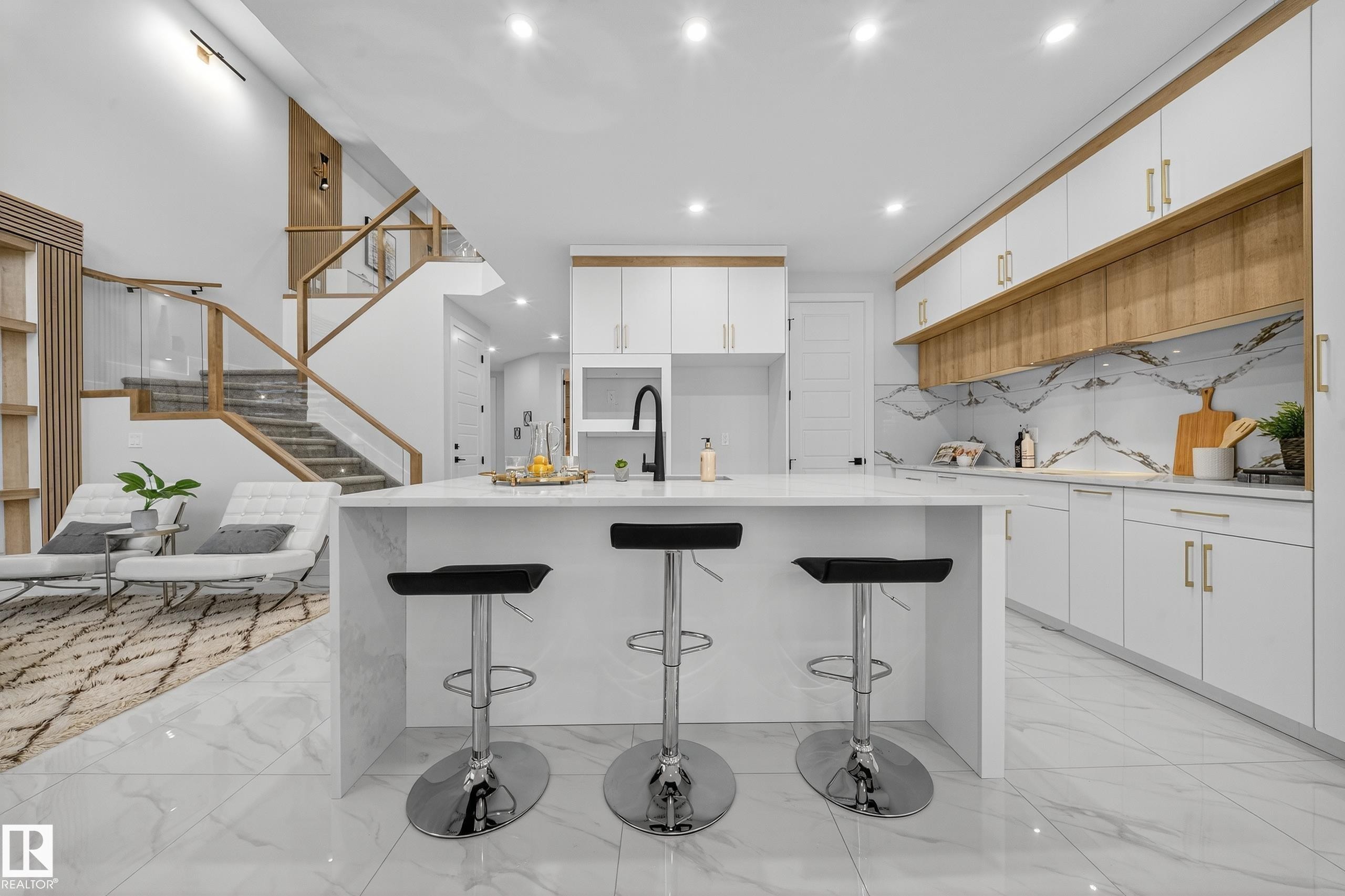 Contemporary kitchen featuring a large waterfall island with integrated seating, extensive white cabinetry, wood-finish accents, and a monochrome backsplash - 372 Creekside Green, Leduc, AB