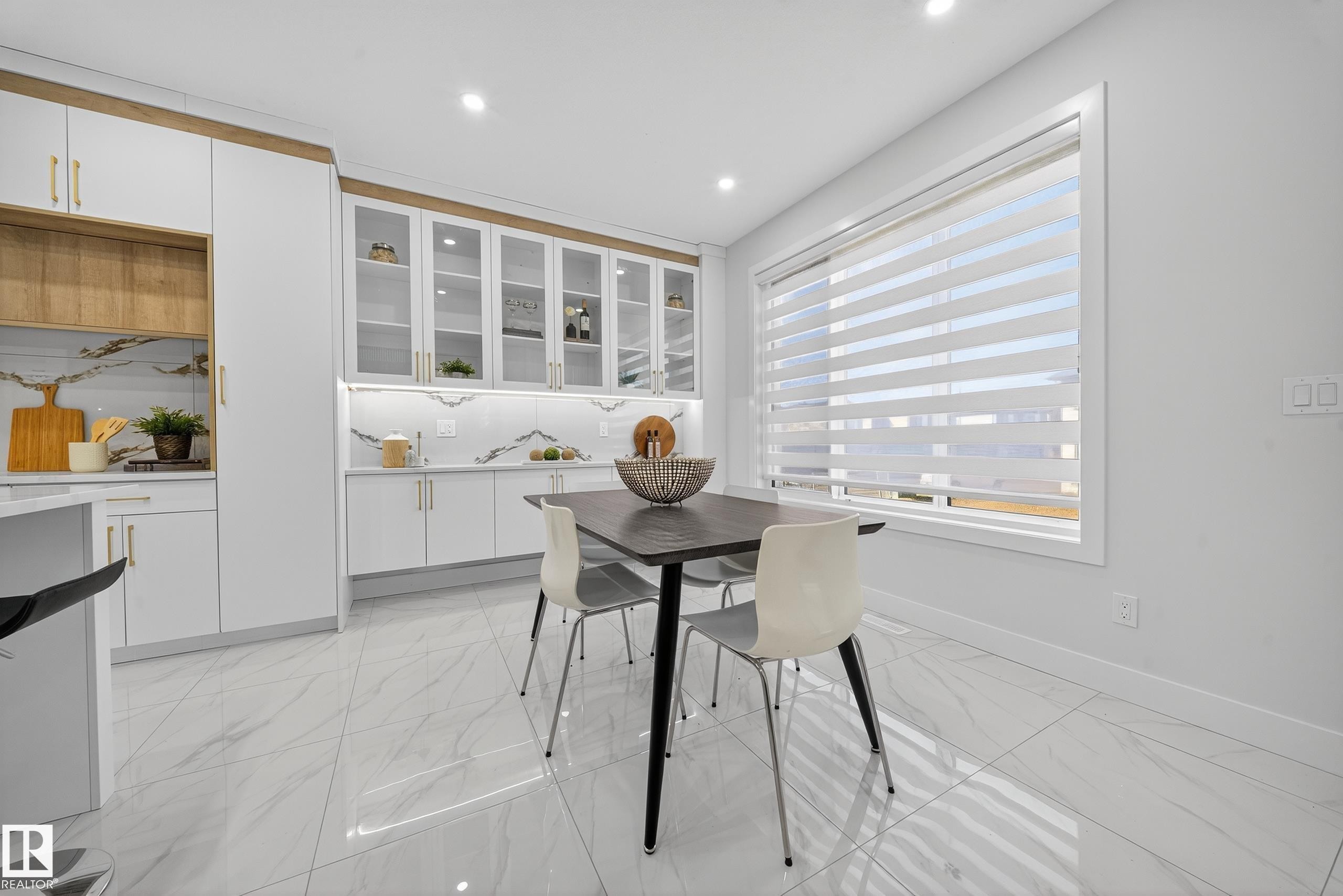 Contemporary kitchen and dining area featuring white cabinetry with gold-tone hardware, wood-finish accents, and a marble-patterned backsplash - 372 Creekside Green, Leduc, AB