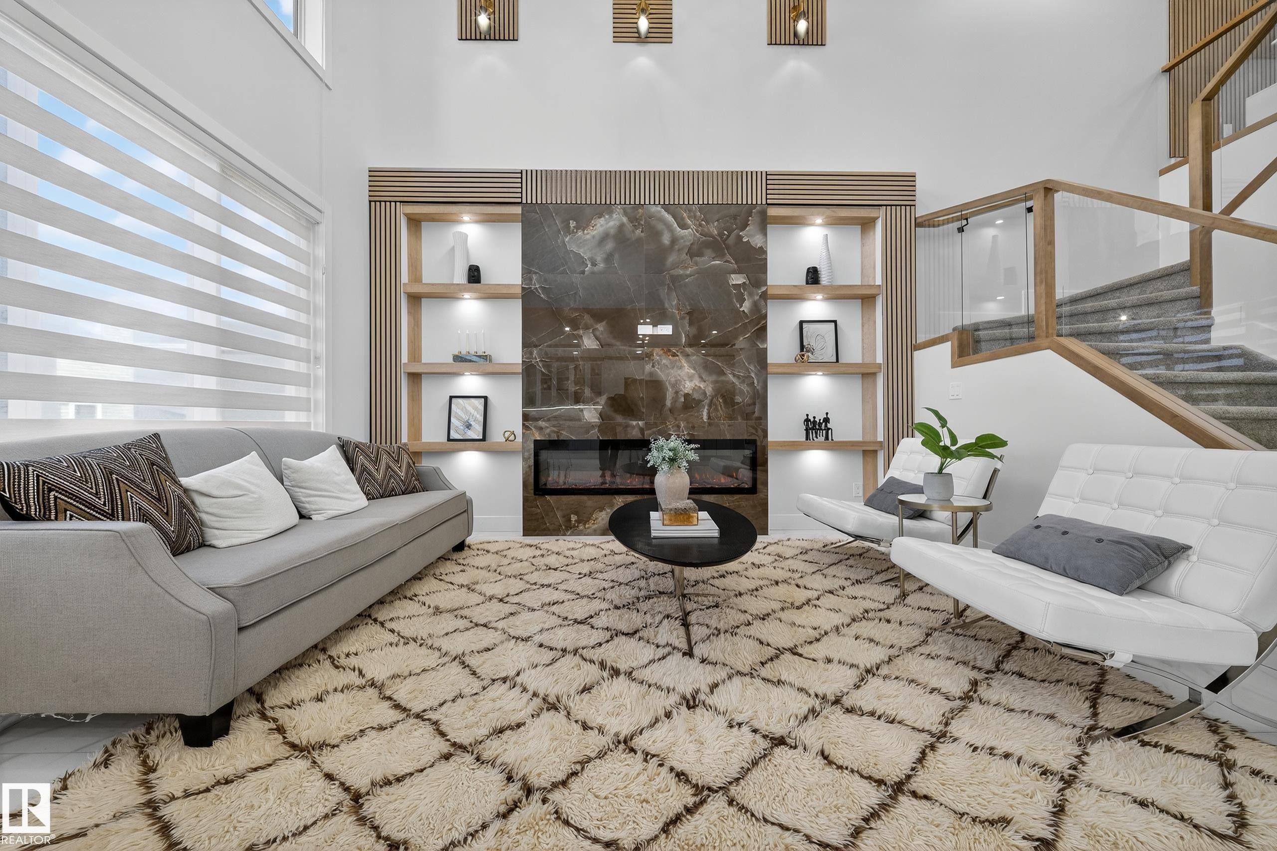 Living area featuring a floor-to-ceiling fireplace with stone surround and built-in shelving, a modern staircase with glass railing and wood handrail, high ceilings, and large windows with horizontal blinds - 372 Creekside Green, Leduc, AB
