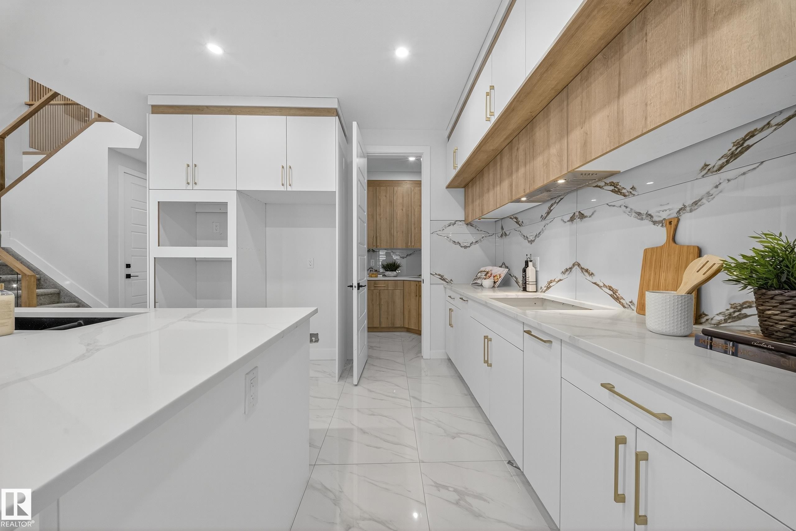 Contemporary kitchen featuring a large island with a waterfall-edge countertop, white cabinetry with gold-toned hardware, a full-height backsplash with a unique veining pattern, wood-finish accent shelving, and integrated lighting - 372 Creekside Green, Leduc, AB
