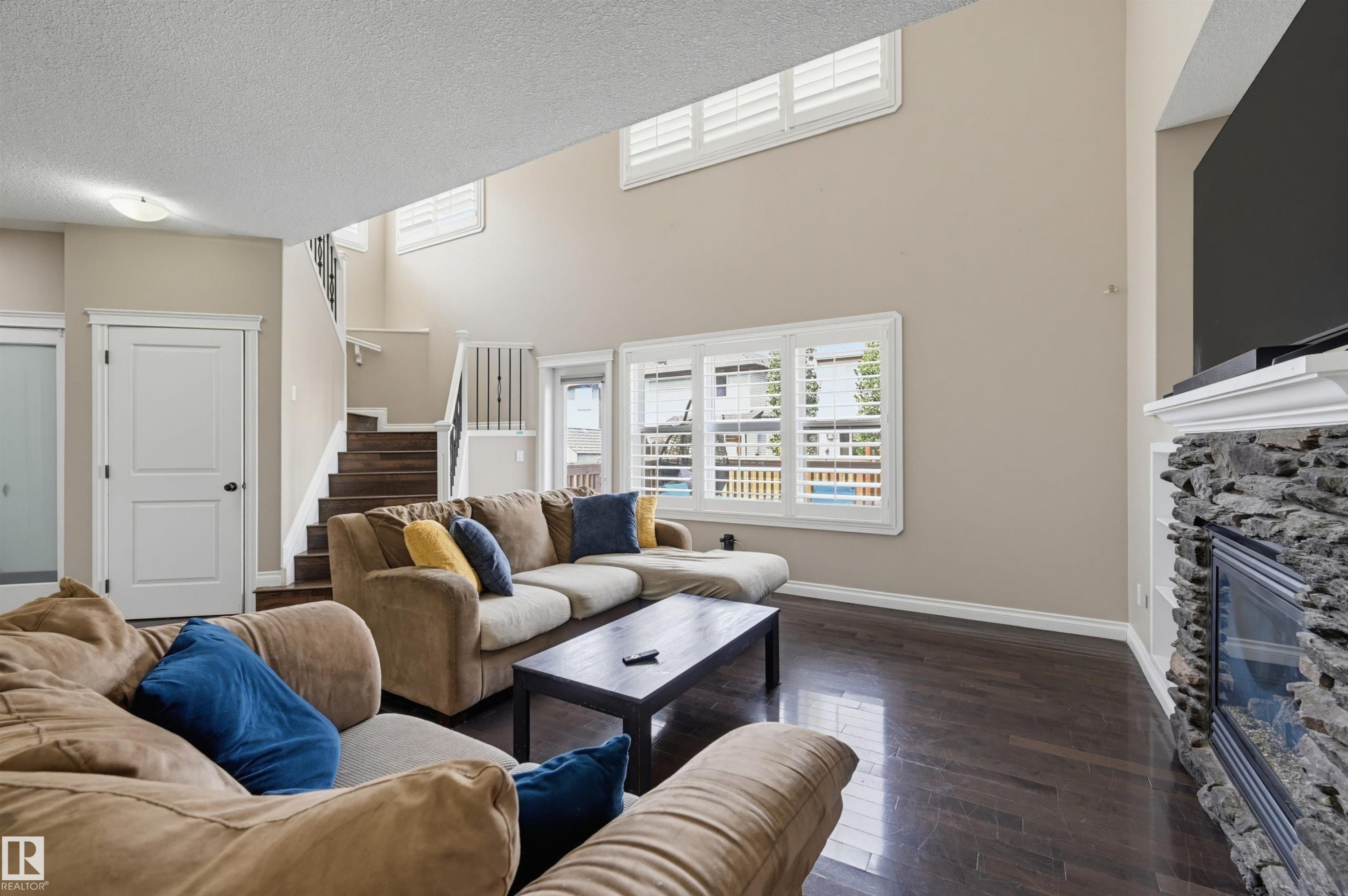 Spacious living area featuring high ceilings, dark wood-finish flooring, a stone-clad fireplace with a white mantel, and multiple windows with plantation shutters - 21928 97 Avenue Nw, Edmonton, AB - Indoor Photo Showing Living Room With Fireplace