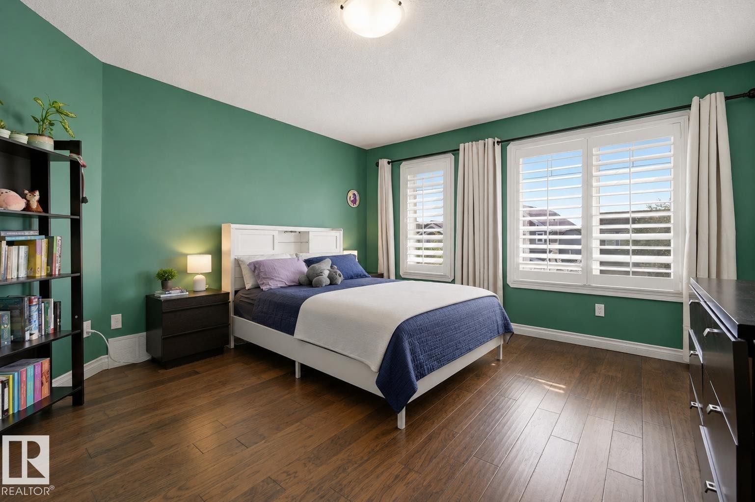 Bedroom featuring rich wood-finish flooring, vibrant green walls, two windows with plantation shutters, white baseboards, and a flush-mount ceiling light fixture - 21928 97 Avenue Nw, Edmonton, AB - Indoor Photo Showing Bedroom