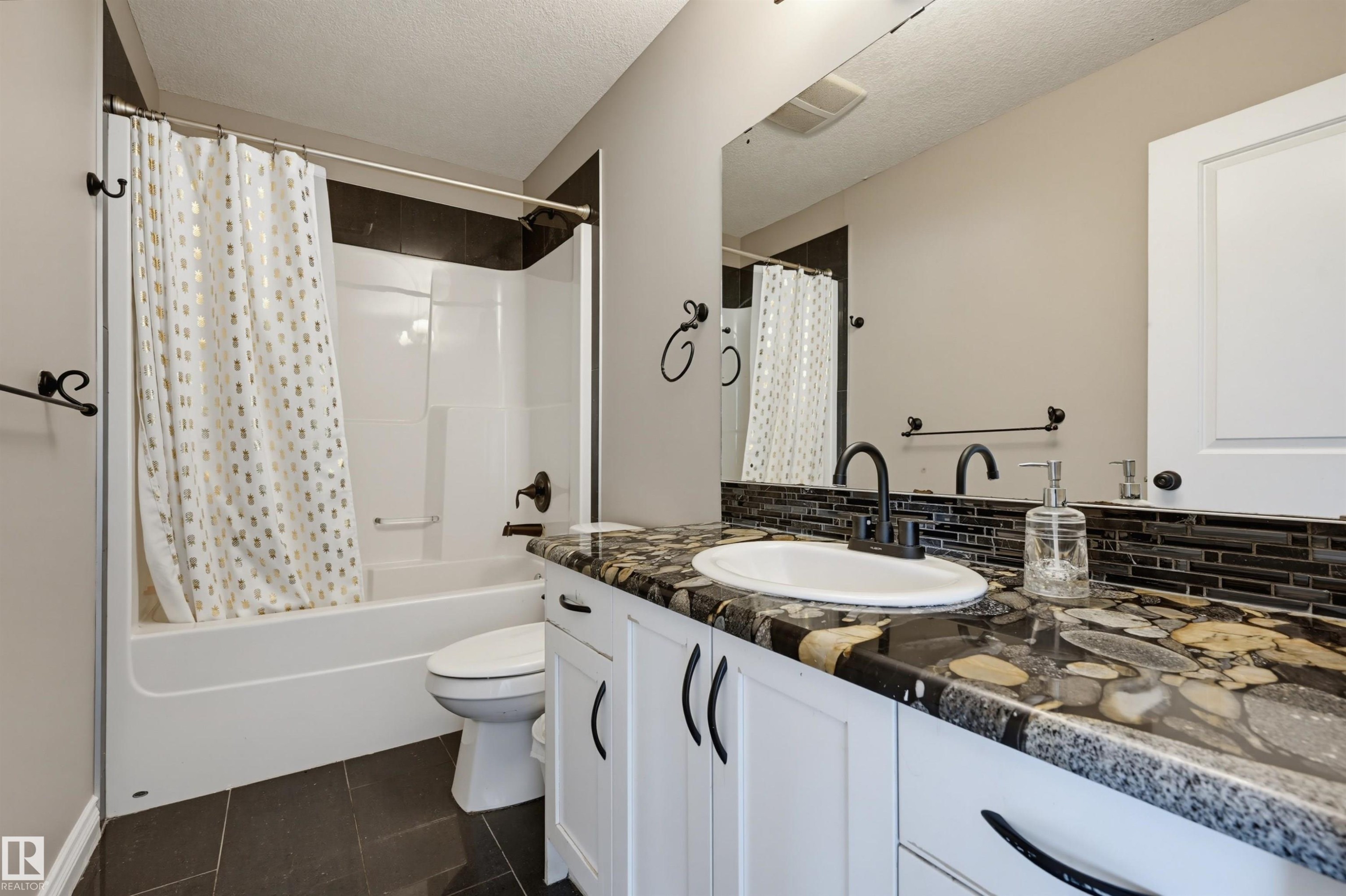 Bathroom featuring a white vanity with black hardware, a unique stone-patterned countertop, a black subway tile backsplash, and a white tub/shower combination with dark bronze fixtures - 21928 97 Avenue Nw, Edmonton, AB - Indoor Photo Showing Bathroom