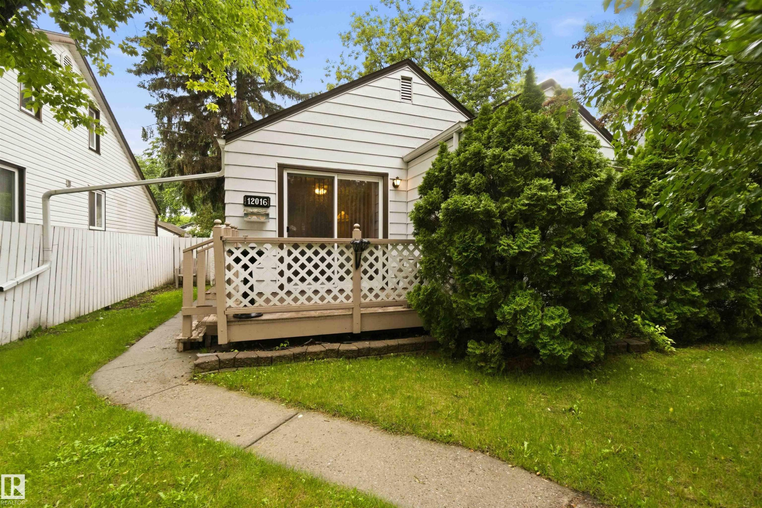 White siding exterior featuring a front deck with lattice railing, mature shrubs, a concrete pathway, and a picket fence - 12016 61 Street, Edmonton, AB