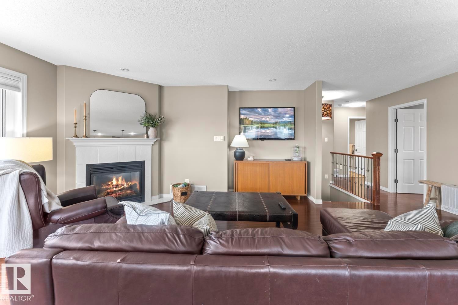 Living area featuring a white mantel fireplace, warm neutral wall tones, wood-finish flooring, a white window blind, and a wood railing with black metal spindles - 2602 Delta Road, Cold Lake, AB