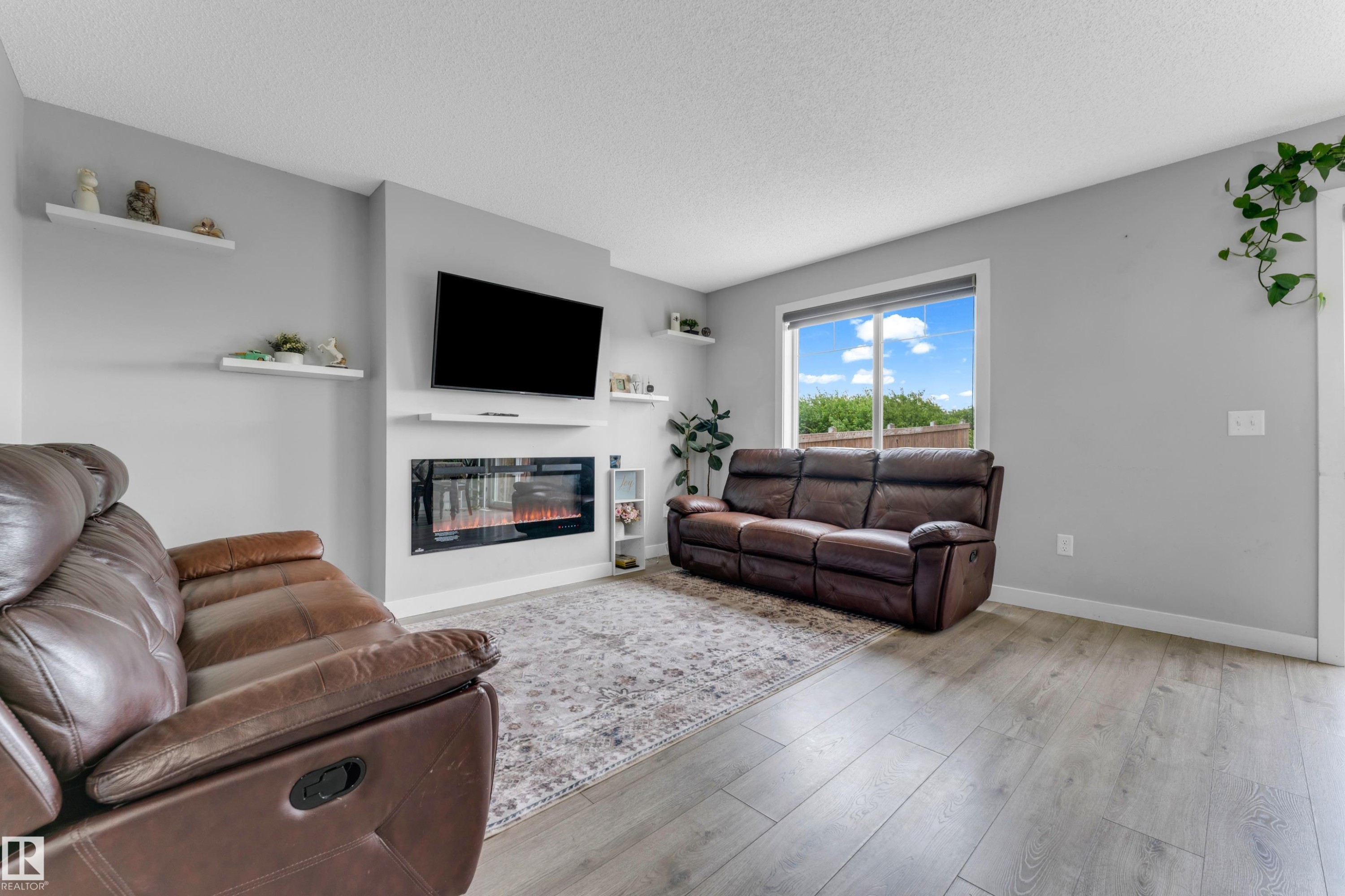 Living room featuring light wood-finish flooring, a contemporary electric fireplace with built-in shelving, a large picture window, and light gray wall paint - 1750 27 Street, Edmonton, AB