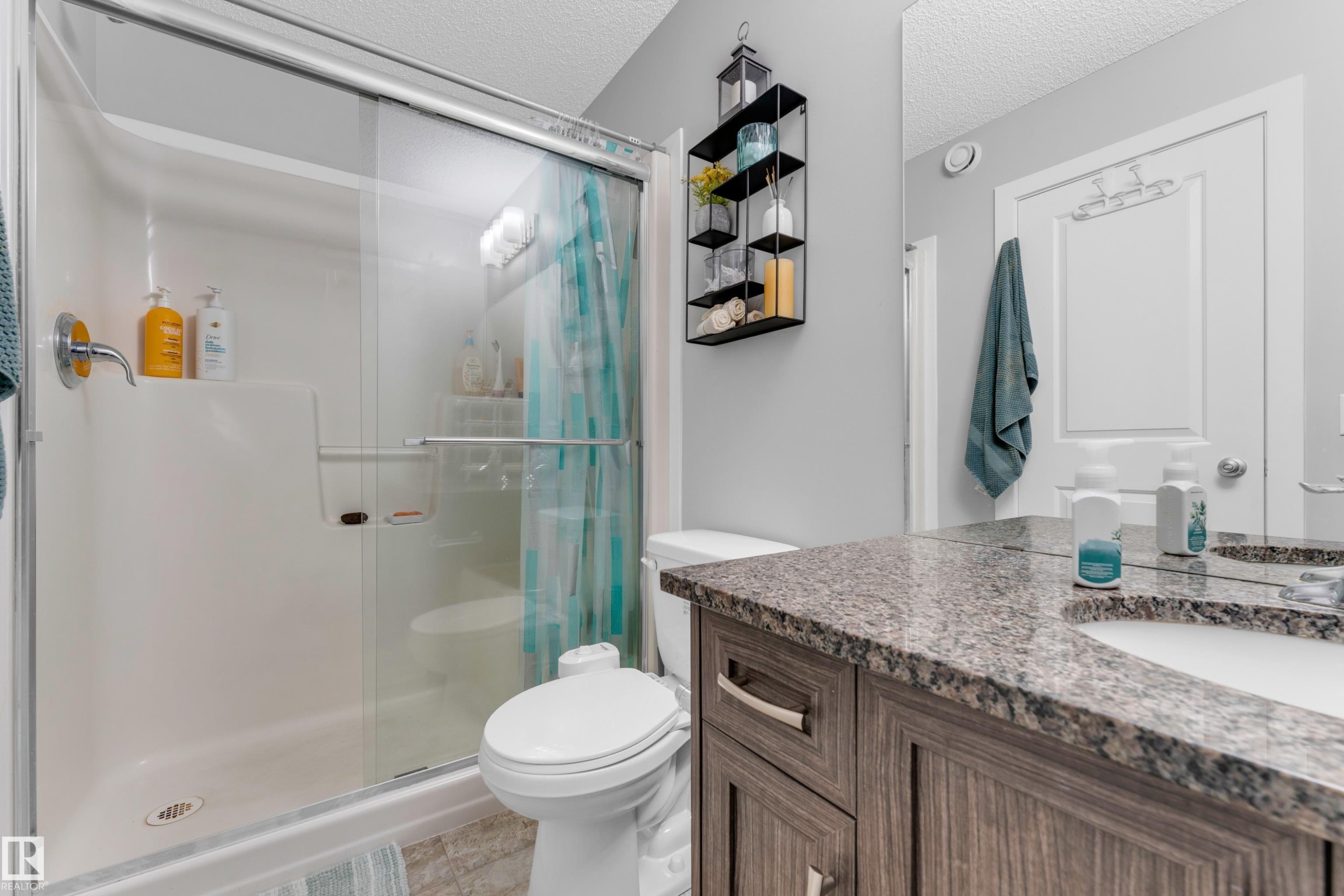 Bathroom featuring a glass-enclosed shower with a built-in shelf, a toilet, and a vanity with a stone-look countertop and an integrated sink - 1750 27 Street, Edmonton, AB