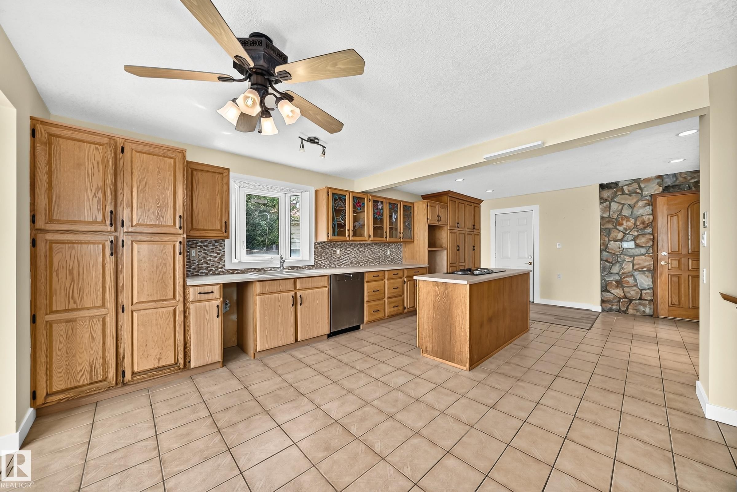 Spacious kitchen featuring extensive wood cabinetry, a central island with a cooktop, a tile backsplash, and tile flooring - 18636 71 Avenue, Edmonton, AB
