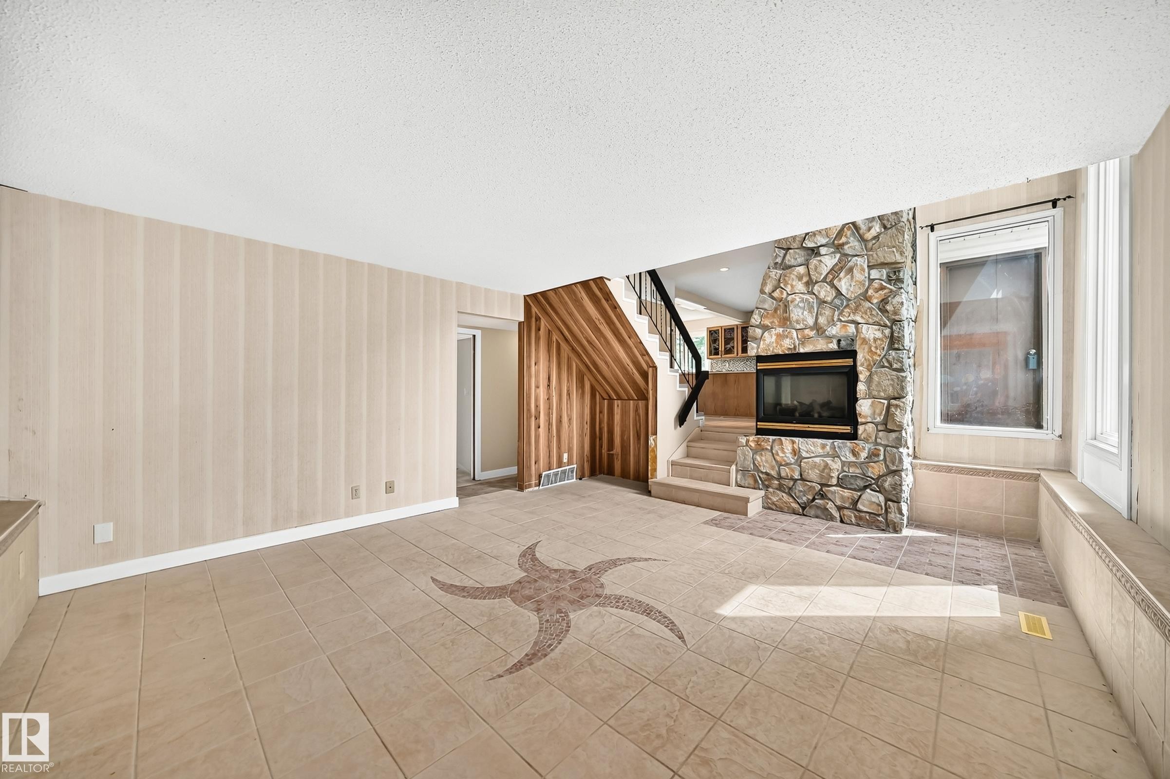 Spacious interior featuring a decorative tile inlay, a stone-clad fireplace with an insert, tiled flooring, wood-finish paneling, and an interior staircase with dark railings - 18636 71 Avenue, Edmonton, AB