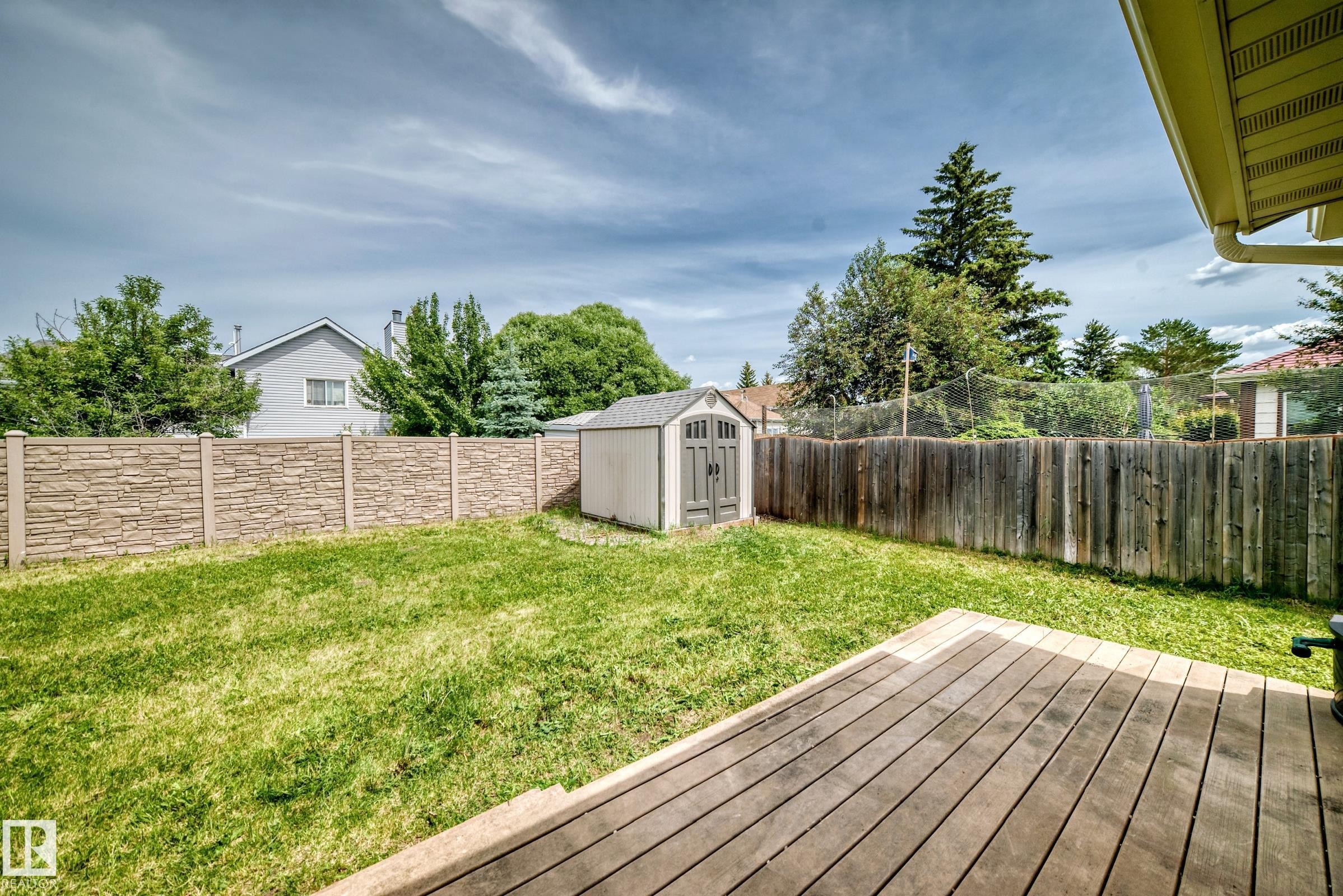 Expansive green lawn featuring a wooden deck, a storage shed, and a stone-look fence - 18636 71 Avenue, Edmonton, AB
