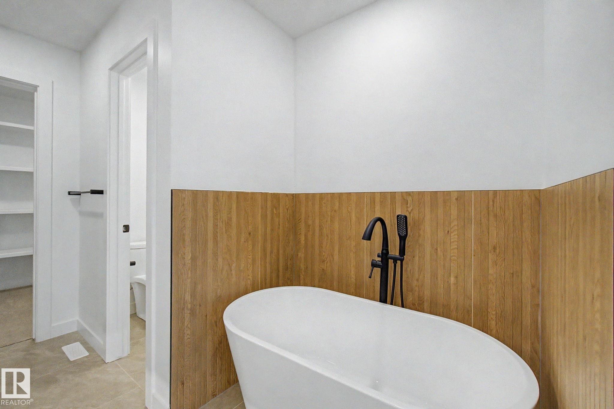Freestanding soaking tub with a matte black floor-mounted faucet, complemented by wood-finish wainscoting and light-colored tile flooring - 4 Willow Heights Drive, Stony Plain, AB