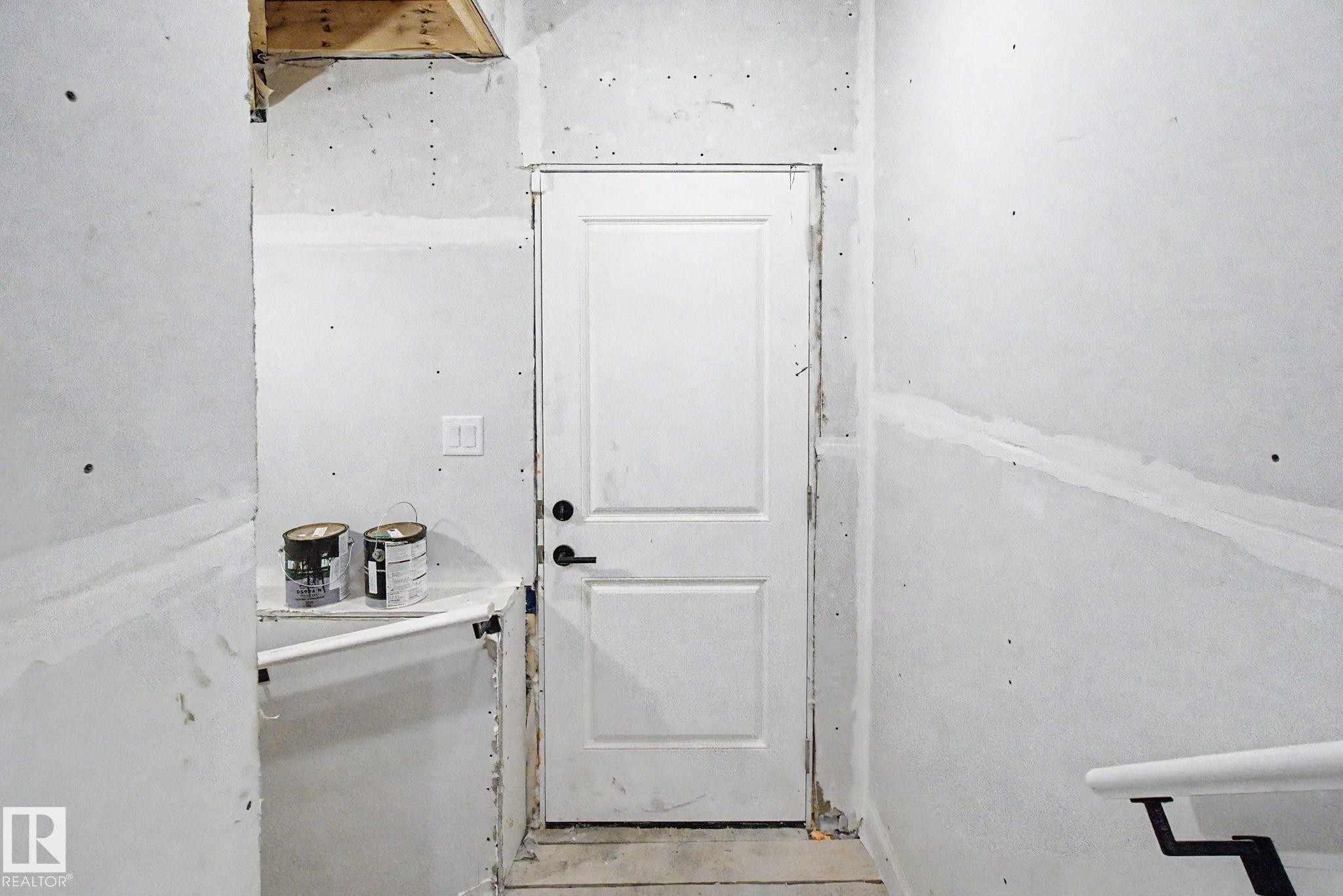 Interior space featuring a white two-panel door with black hardware, unfinished drywall, exposed ceiling joists, and a partial white handrail - 4 Willow Heights Drive, Stony Plain, AB