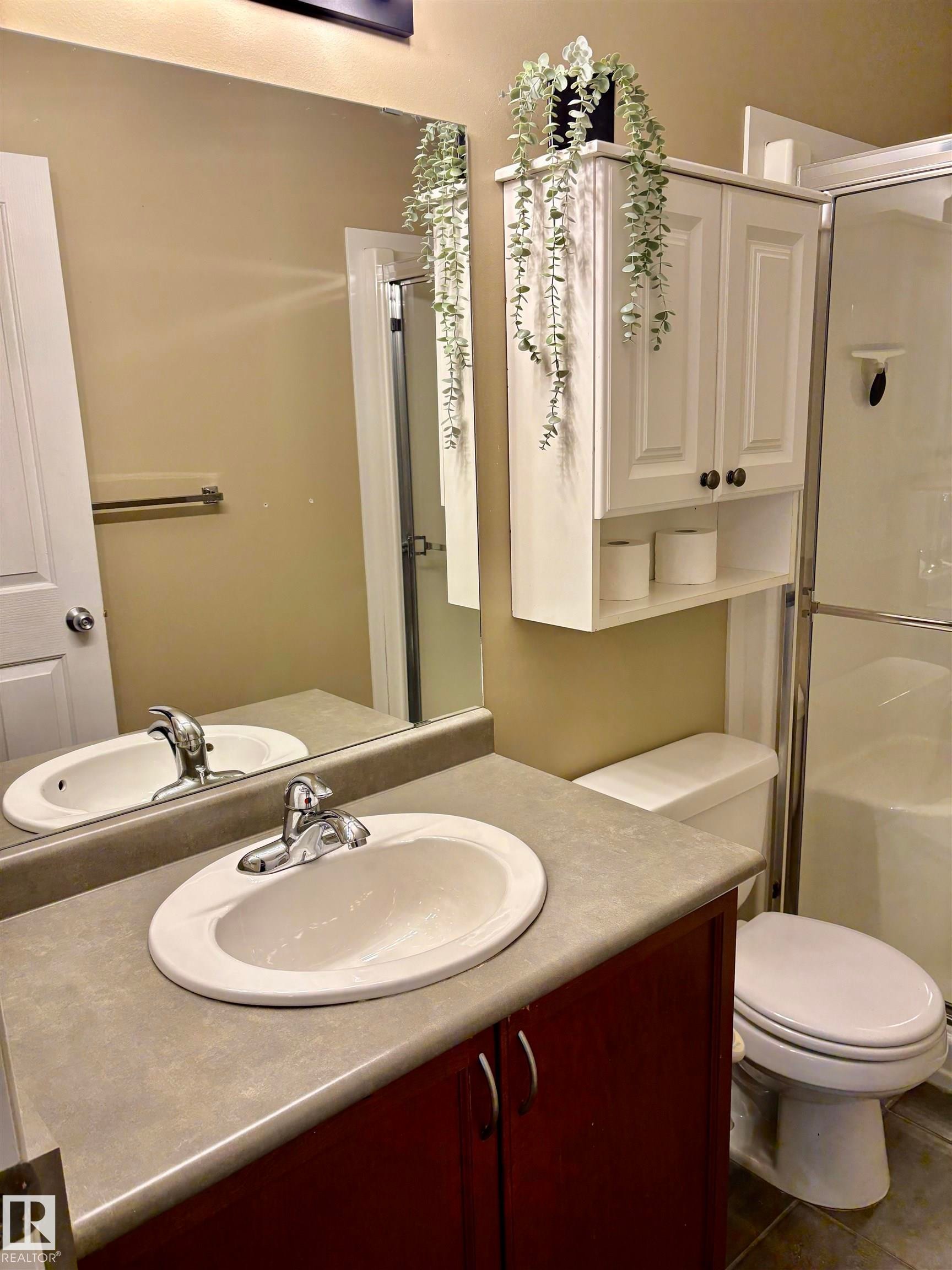 Bathroom featuring a vanity with a dark wood-finish cabinet and integrated sink, a large mirror, white wall-mounted cabinetry, a toilet, and a stand-up shower with a glass enclosure - 184 1804 70 Street, Edmonton, AB