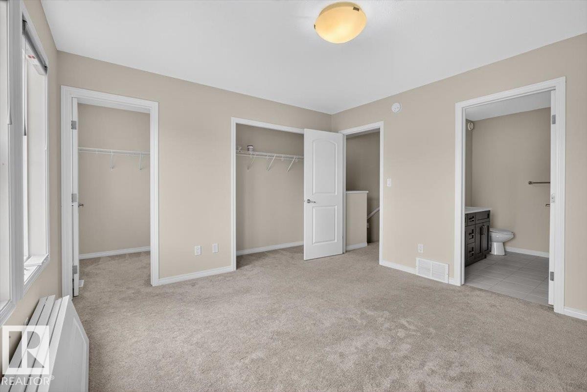 Carpeted bedroom featuring two closet spaces, a window with a radiator, and direct access to a bathroom with a dark wood vanity and tiled flooring - 2144 51 Street, Edmonton, AB