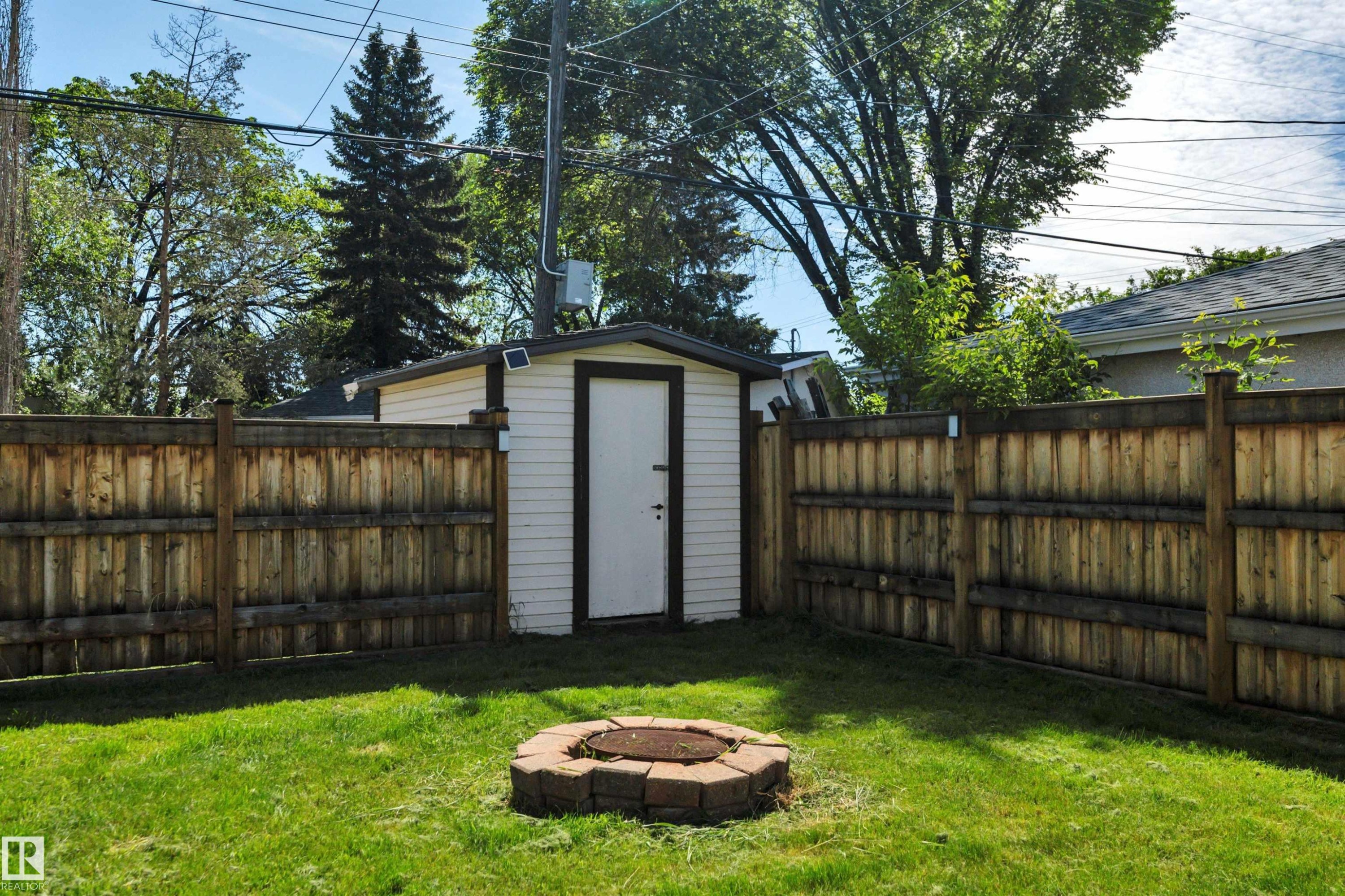 Expansive backyard featuring a manicured lawn, a circular paver fire pit, a wood privacy fence, and a white storage shed with a dark trim - 9835 74 Street, Edmonton, AB