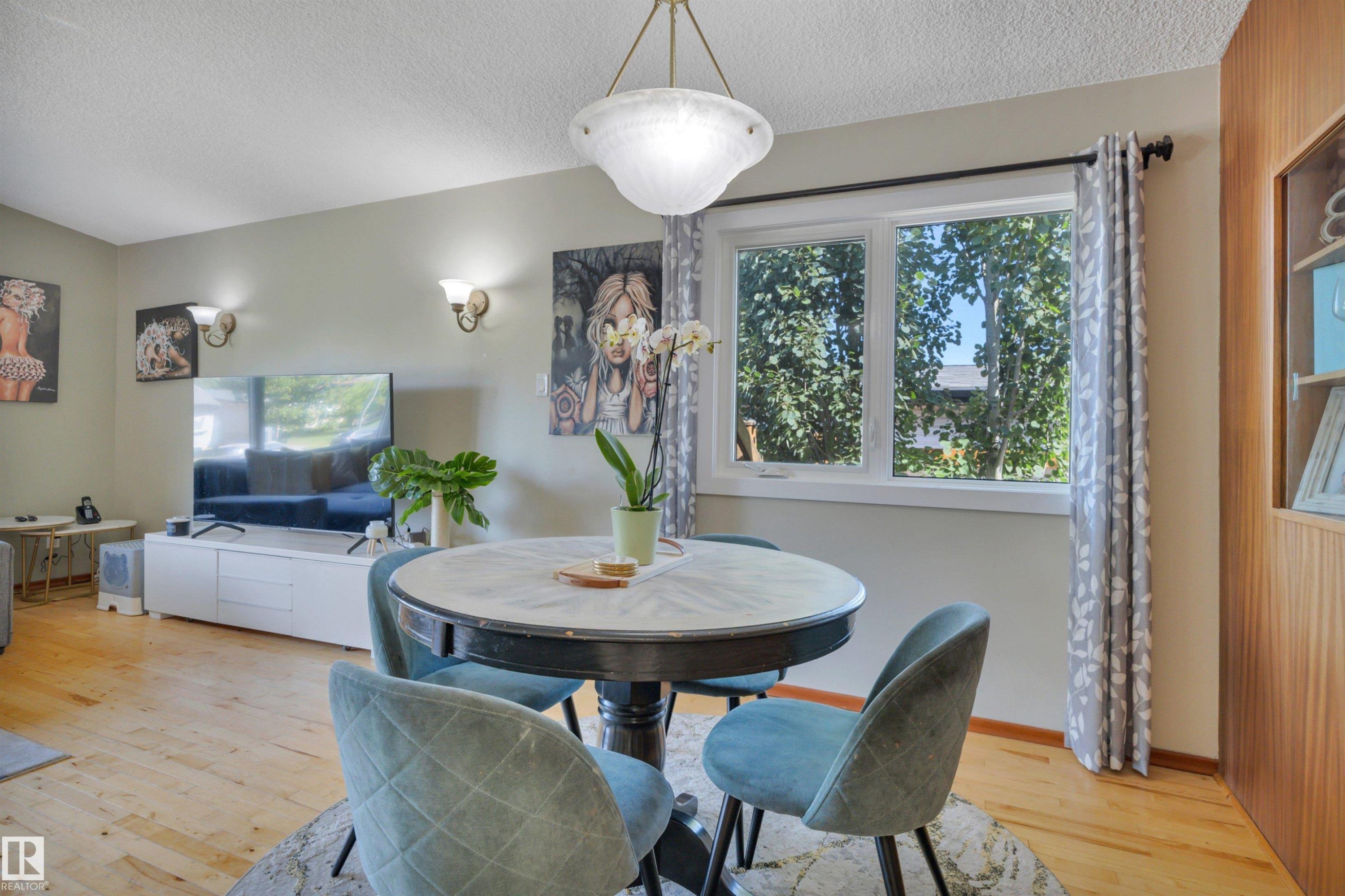 Dining area featuring a central chandelier, wood-finish flooring, a double window with exterior tree coverage, and a built-in vertical wood-paneled display cabinet - 9835 74 Street, Edmonton, AB