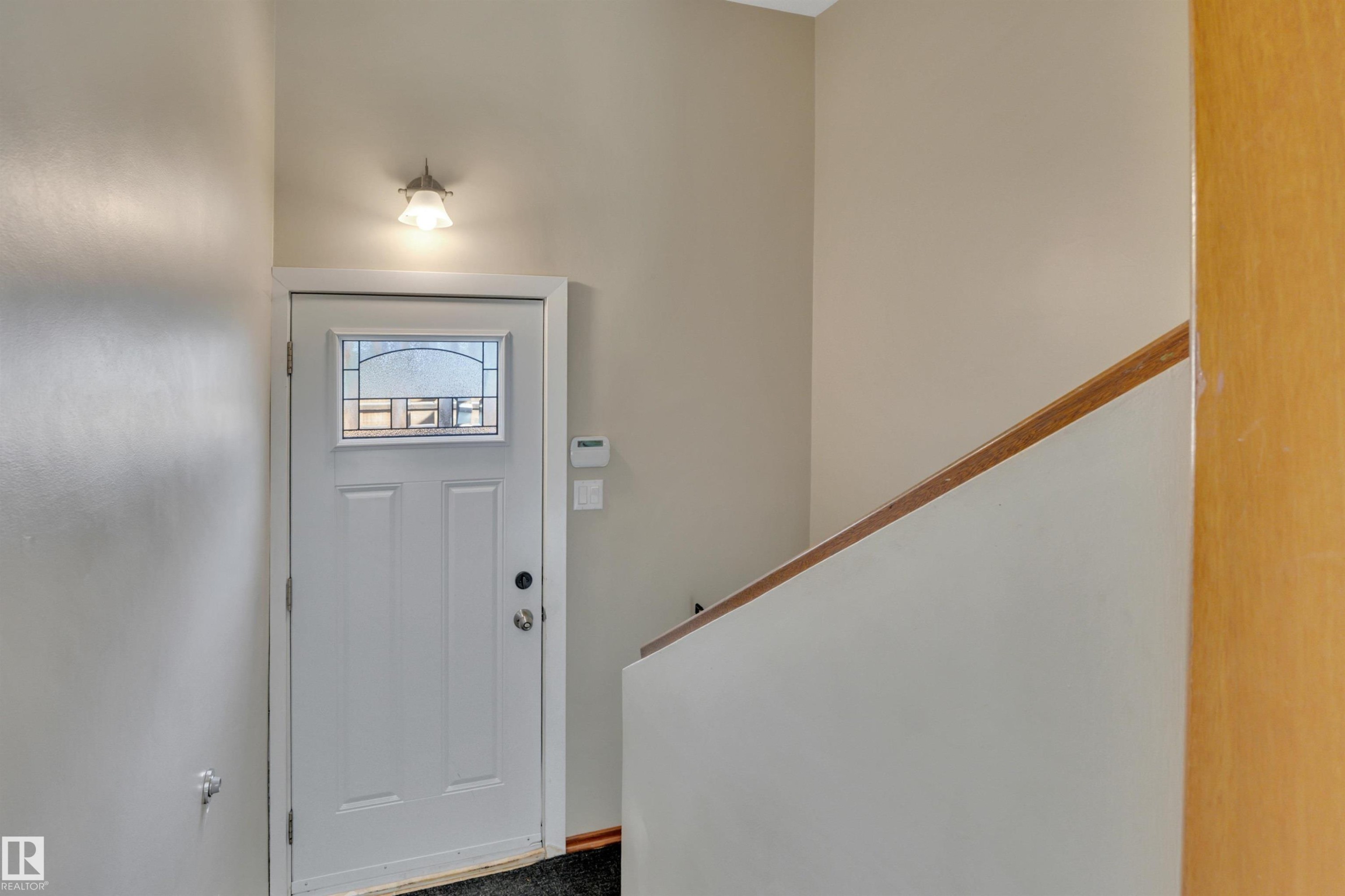 Entryway featuring a white paneled door with decorative glass, a wall-mounted light fixture, and a light-toned wood handrail - 9835 74 Street, Edmonton, AB