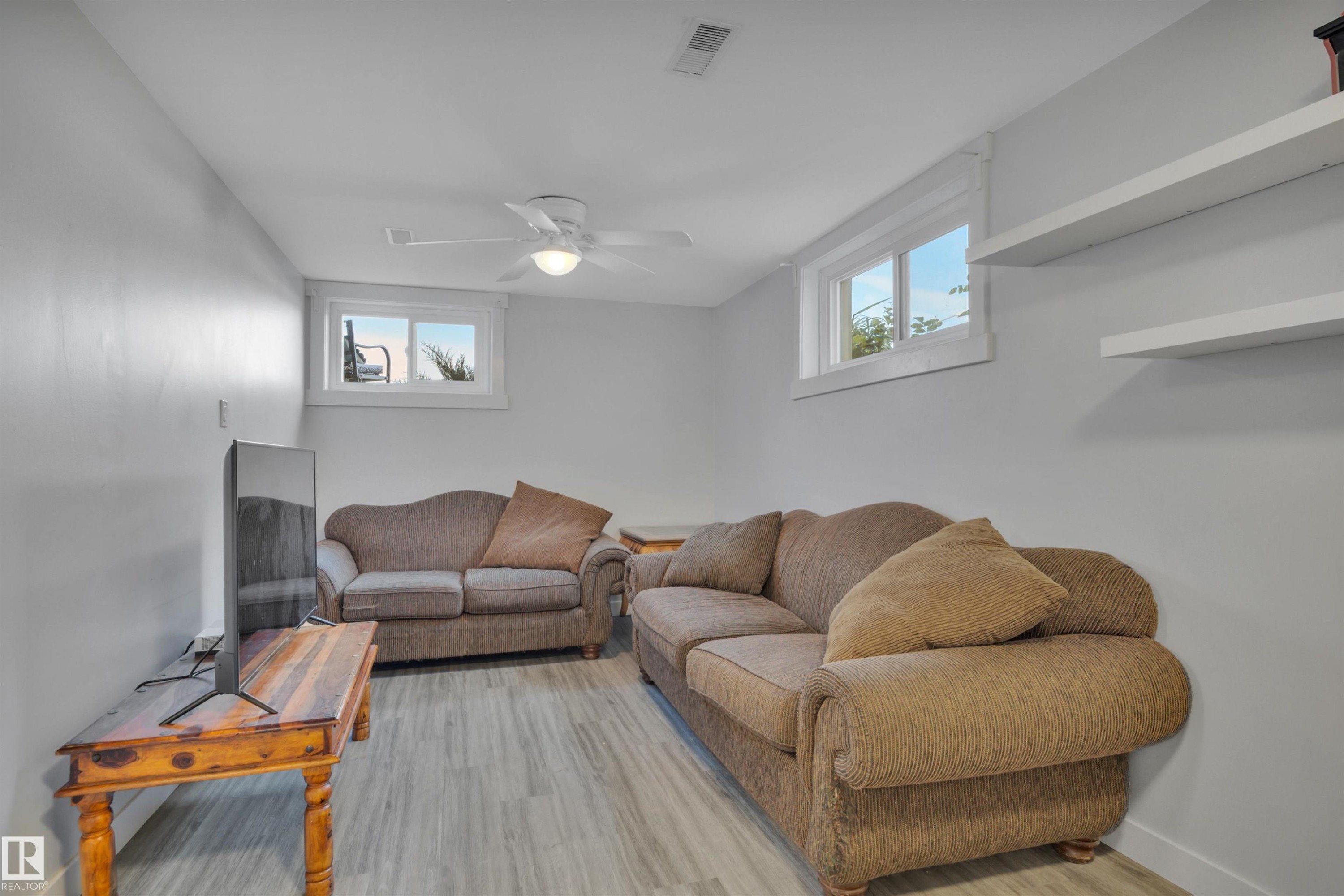 Finished lower level space featuring wood-finish flooring, light gray walls, a ceiling fan, two windows, and two wall-mounted shelves - 9835 74 Street, Edmonton, AB