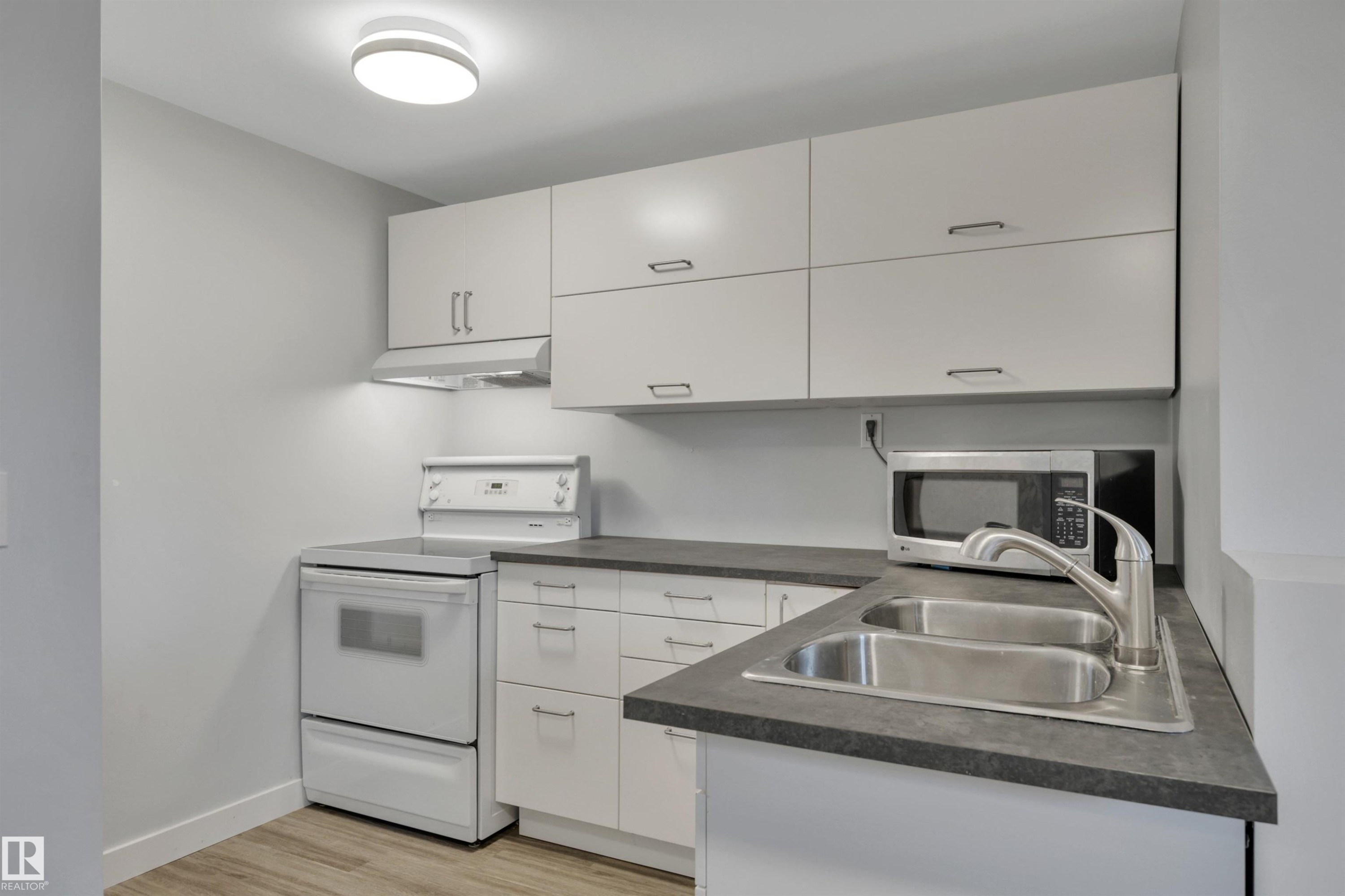 Kitchen featuring light-toned cabinetry with brushed nickel hardware, dark gray countertops, a stainless steel double-basin sink with a gooseneck faucet, a white electric range, and wood-finish flooring - 9835 74 Street, Edmonton, AB