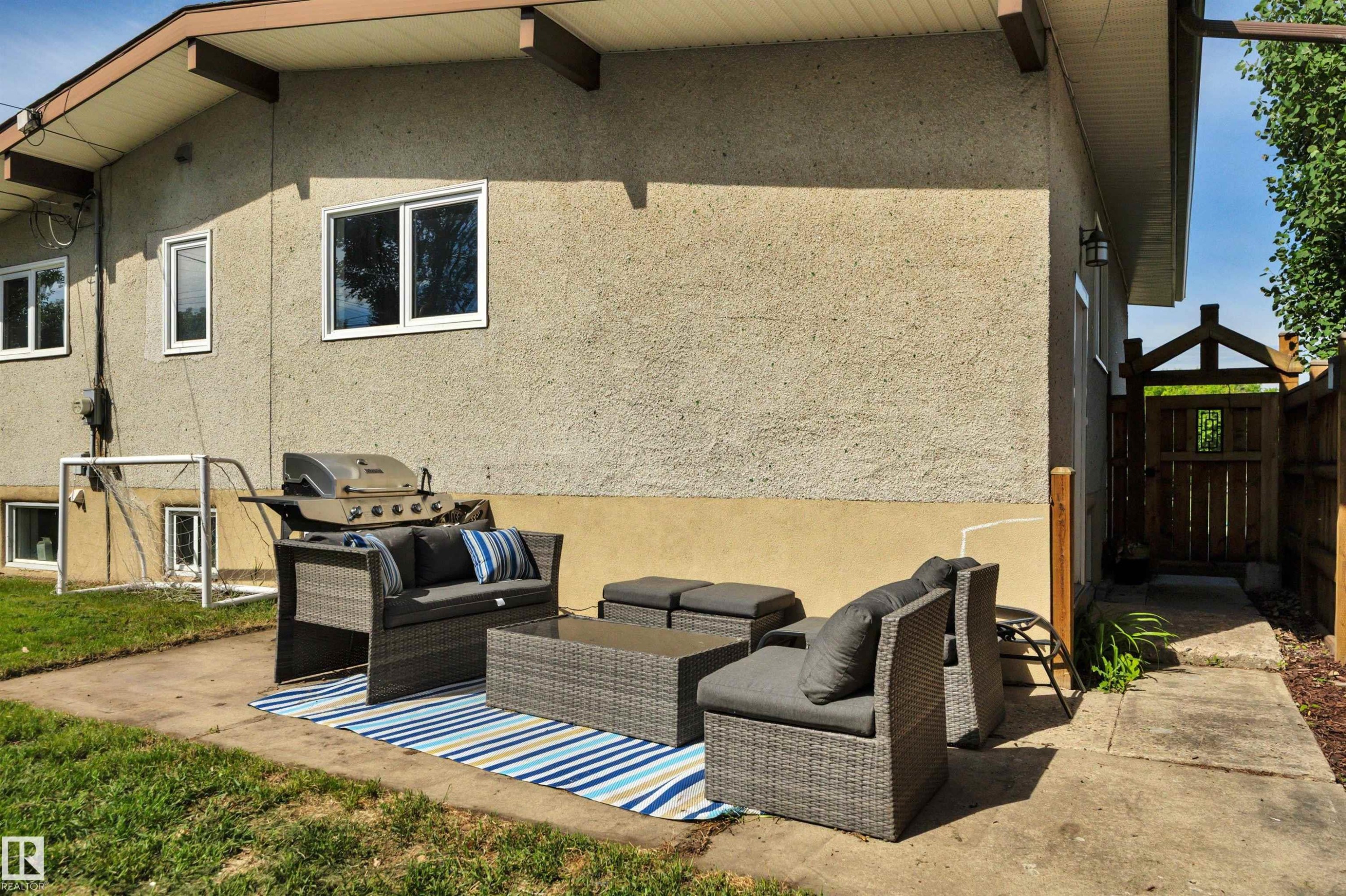 Exterior patio area featuring a concrete pad, stucco siding, multiple windows, a decorative wooden gate, and a privacy fence - 9835 74 Street, Edmonton, AB