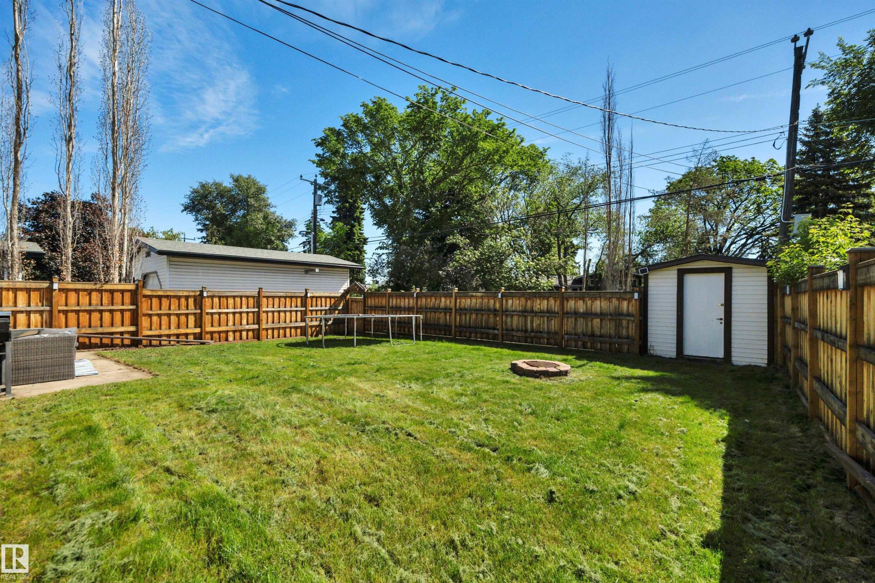 Expansive backyard featuring a manicured lawn, full wood fencing, a dedicated fire pit area, a white storage shed, and a concrete patio pad - 9835 74 Street, Edmonton, AB