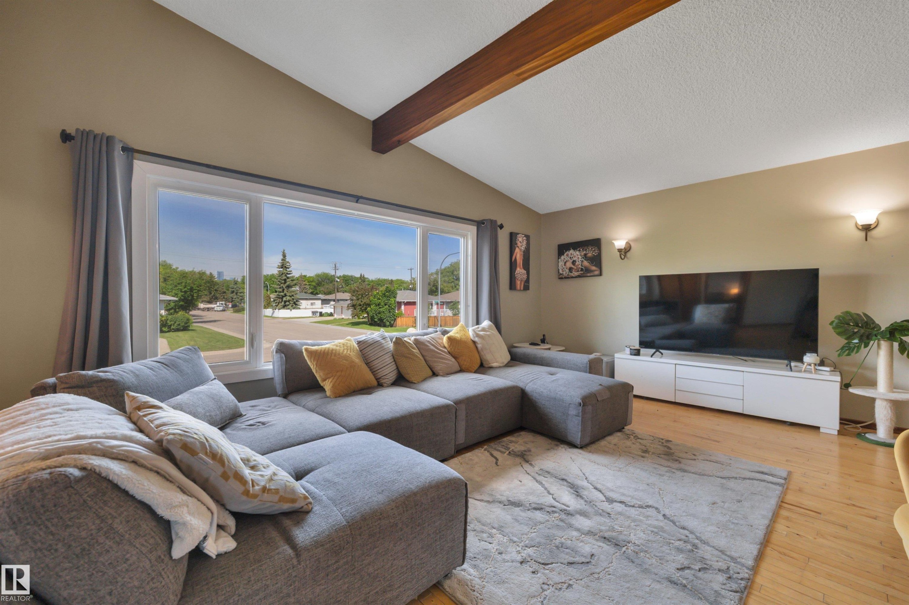 Living area featuring a large picture window, vaulted ceiling with exposed wood beams, wood-finish flooring, and wall-mounted sconce lighting - 9835 74 Street, Edmonton, AB