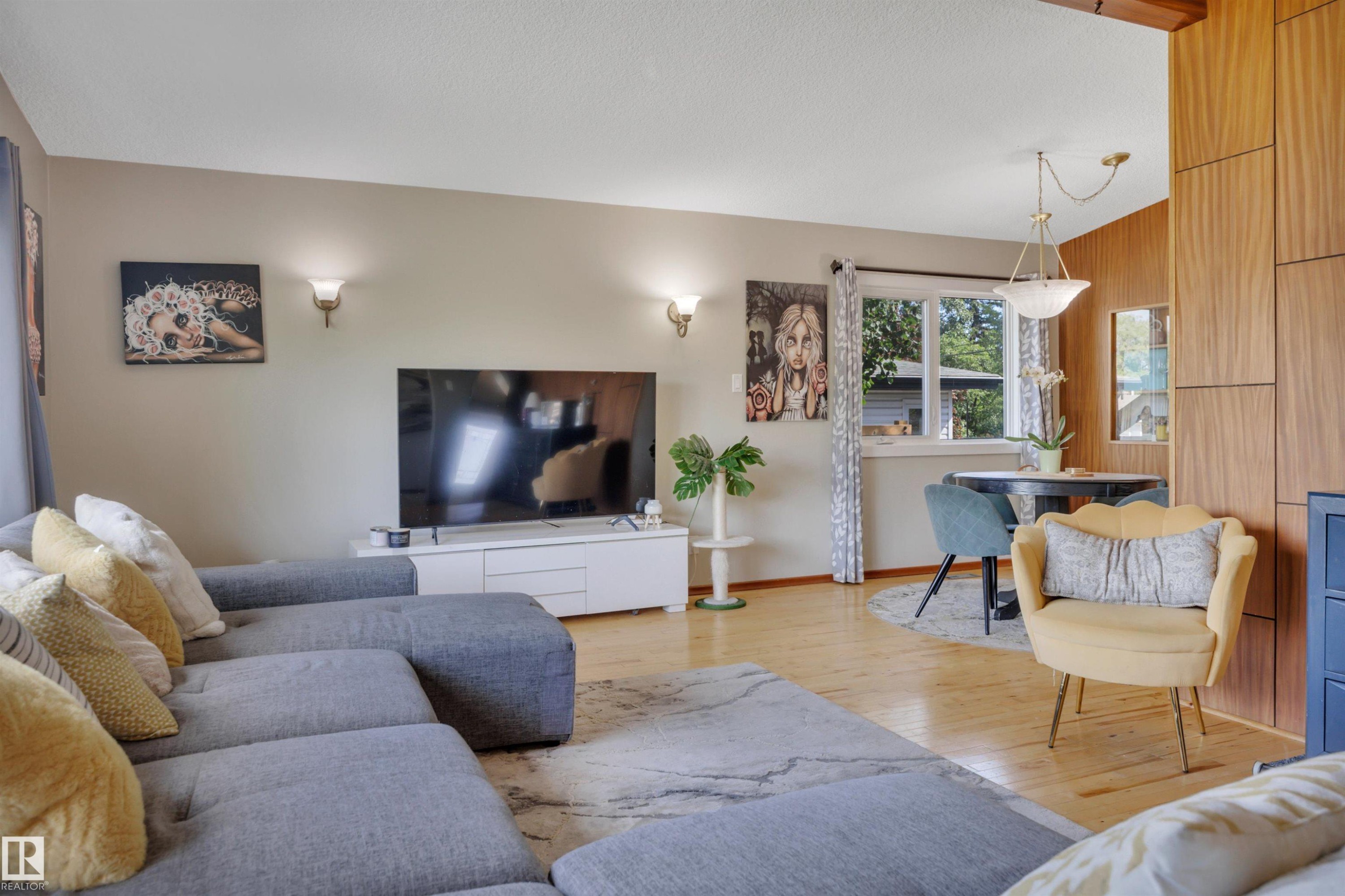Open-concept living area featuring blonde wood-finish flooring, a wood paneled accent wall, a dining area with a circular table, and a tiered chandelier - 9835 74 Street, Edmonton, AB