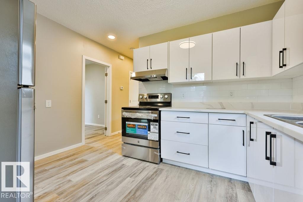 Kitchen featuring sleek white cabinetry with black hardware, stainless steel appliances, a white subway tile backsplash, and light-toned wood-finish flooring - 11241 88 Street, Edmonton, AB