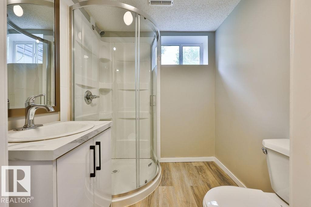 Bathroom featuring a corner shower with curved glass enclosure, a vanity with a white countertop and basin sink, a wall-mounted mirror, and wood-finish flooring - 11241 88 Street, Edmonton, AB
