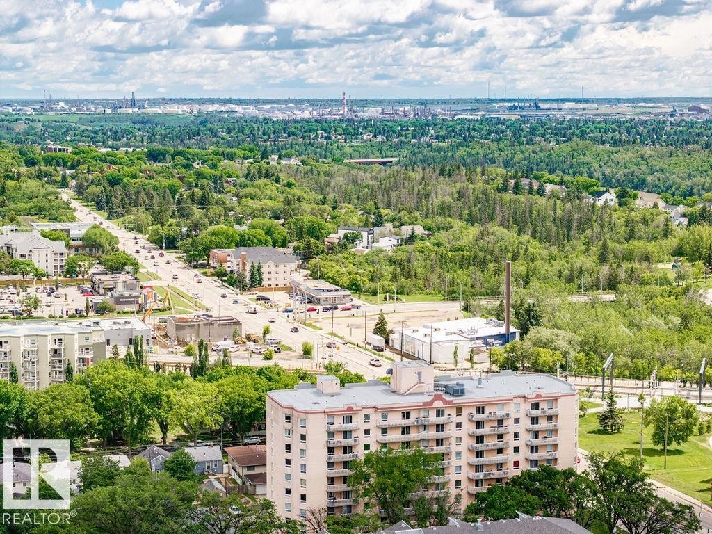 Expansive aerial perspective showcasing a multi-story residential building with multiple balconies, alongside a tree-lined thoroughfare, surrounded by extensive green spaces and distant urban development - 11241 88 Street, Edmonton, AB