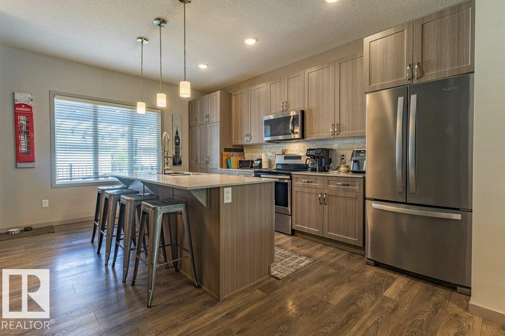 Contemporary kitchen featuring an island with pendant lighting, stainless steel appliances, wood-finish flooring, ample cabinetry, and a window with blinds - 17123 49 Street, Edmonton, AB