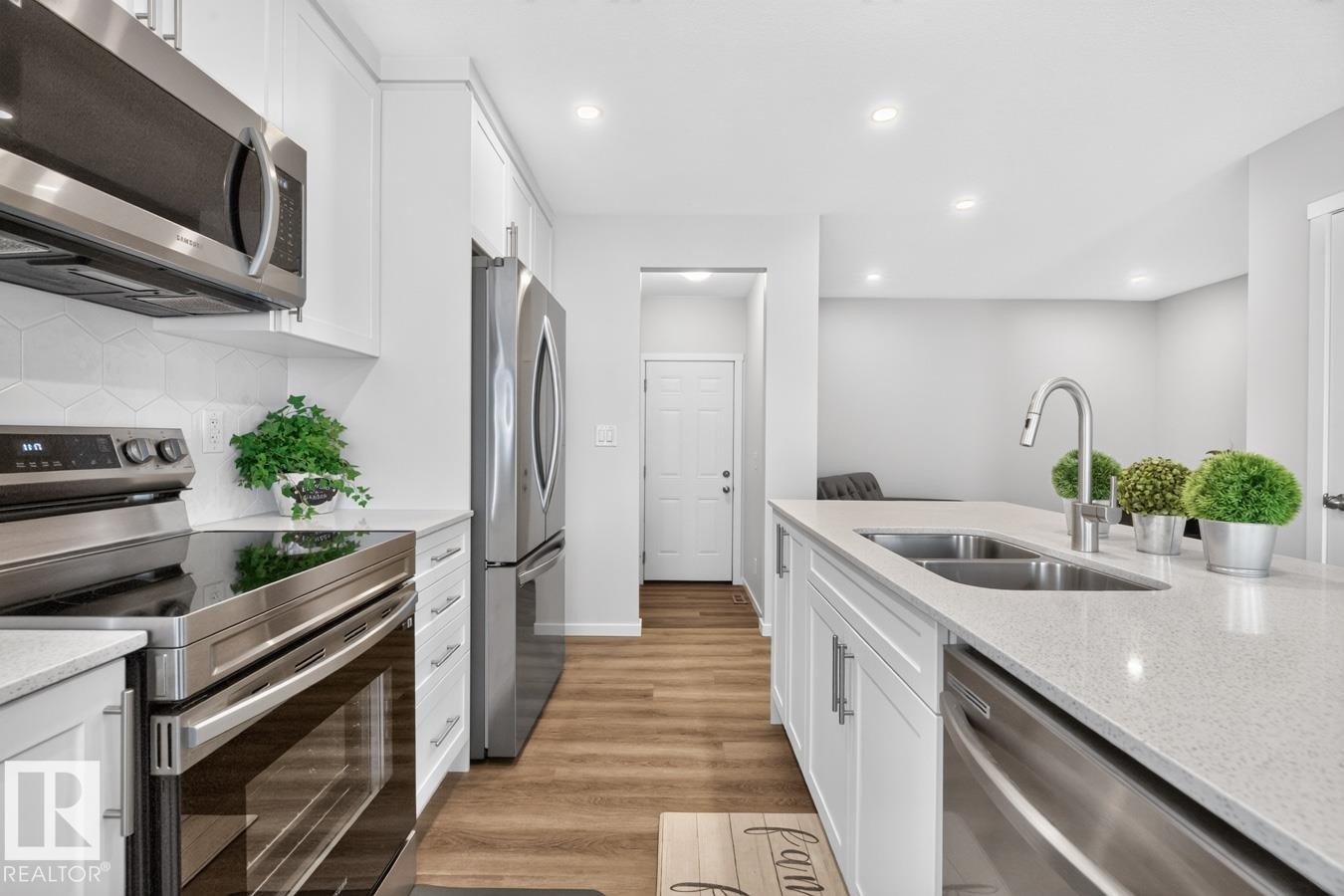 Modern kitchen featuring white shaker cabinetry, stainless steel appliances, a white geometric tile backsplash, wood-finish flooring, and a kitchen island with an undermount double sink - 1022 Hearthstone Boulevard, Sherwood Park, AB