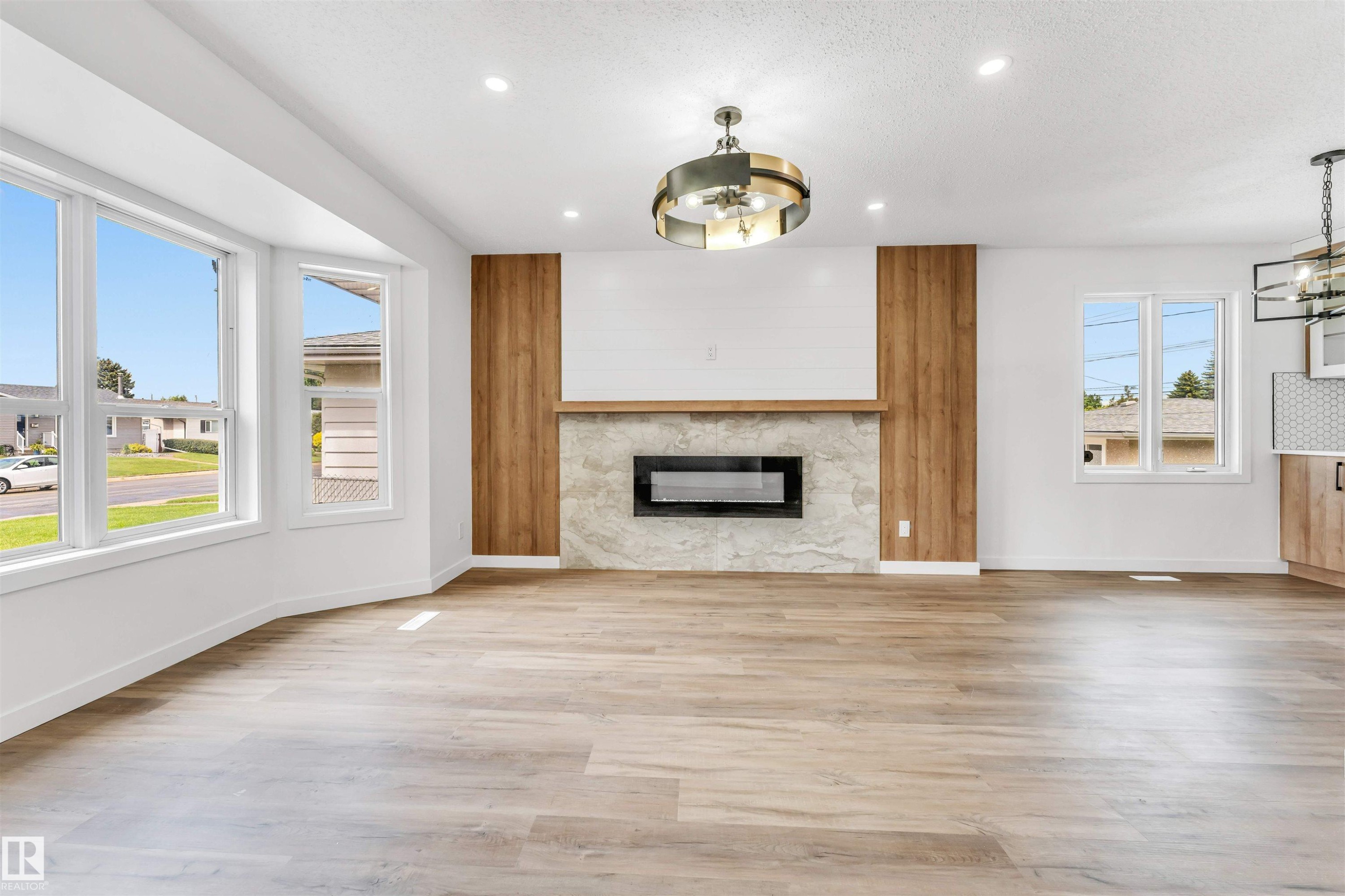 Spacious living area featuring light wood-finish flooring, a modern fireplace with a stone surround and wood paneling, a large bay window, recessed lighting, and a contemporary ceiling fixture - 6404 88 Avenue, Edmonton, AB