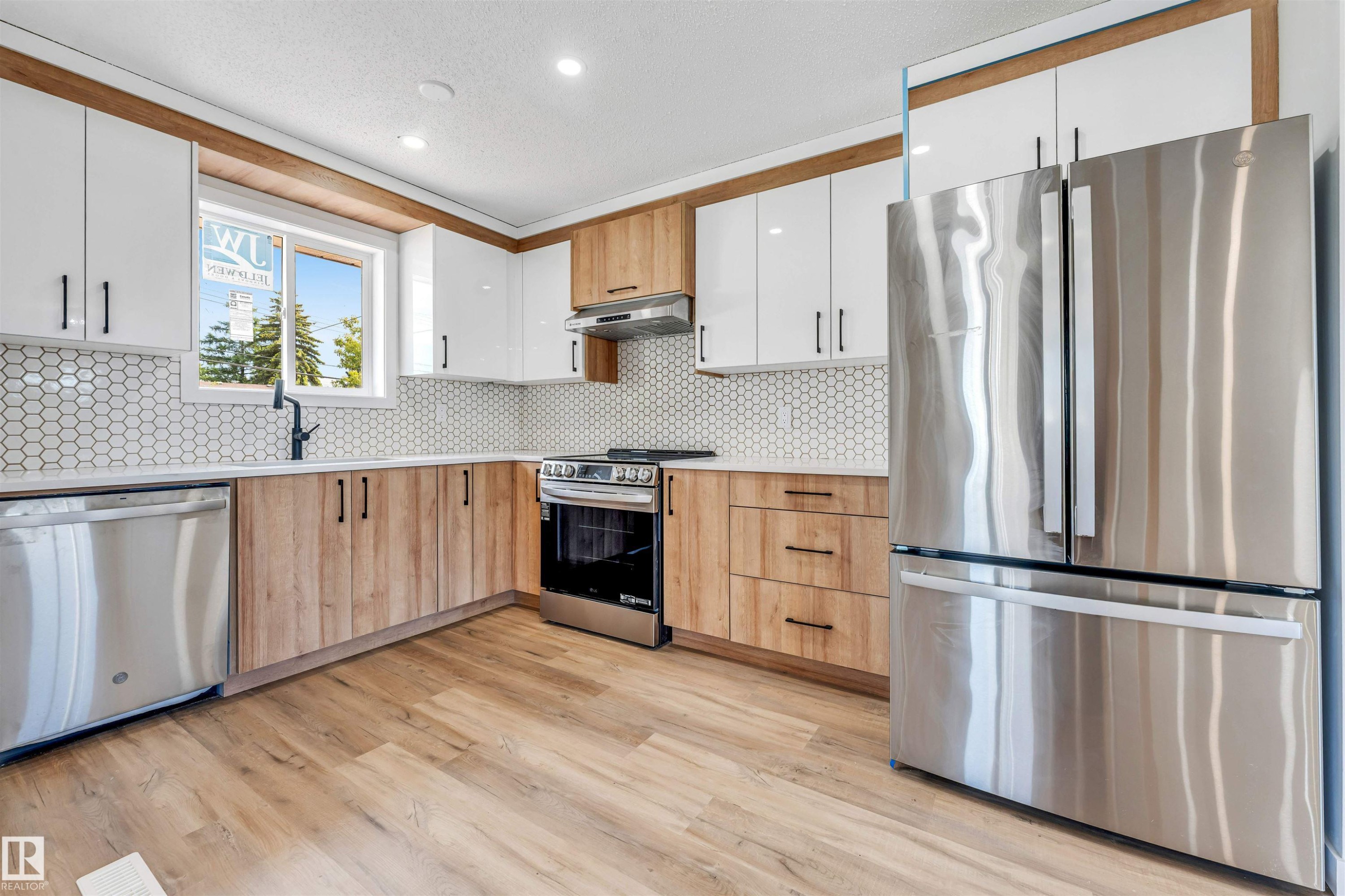 Contemporary kitchen featuring two-tone cabinetry with wood-finish lower cabinets and white upper cabinets, stainless steel appliances including a French door refrigerator, oven range, and dishwasher, a hexagonal tile backsplash, and wood-finish flooring - 6404 88 Avenue, Edmonton, AB