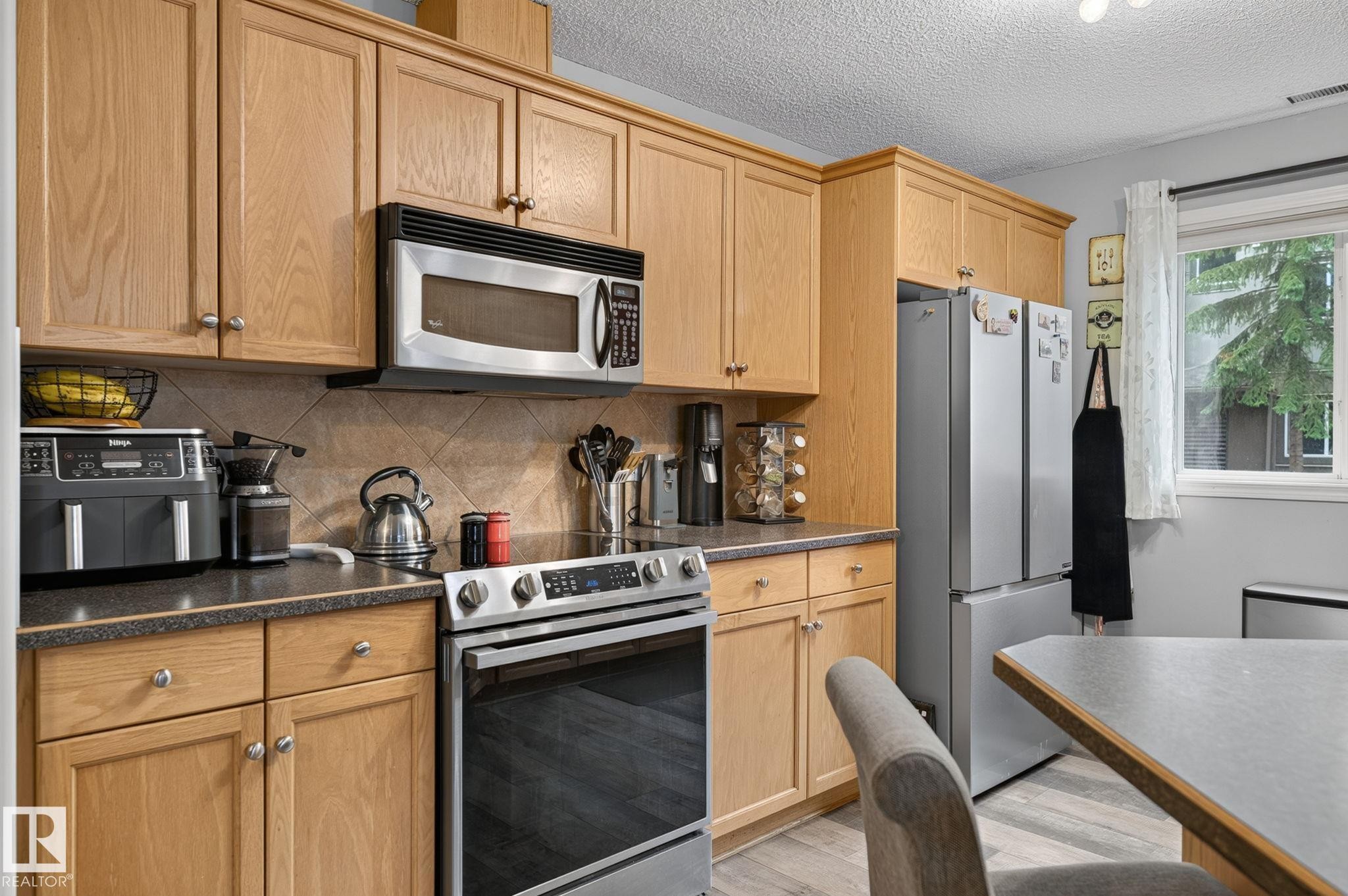 Kitchen featuring light wood cabinetry, stainless steel appliances, dark countertops, and a tiled backsplash - 122 279 Suder Greens Drive, Edmonton, AB