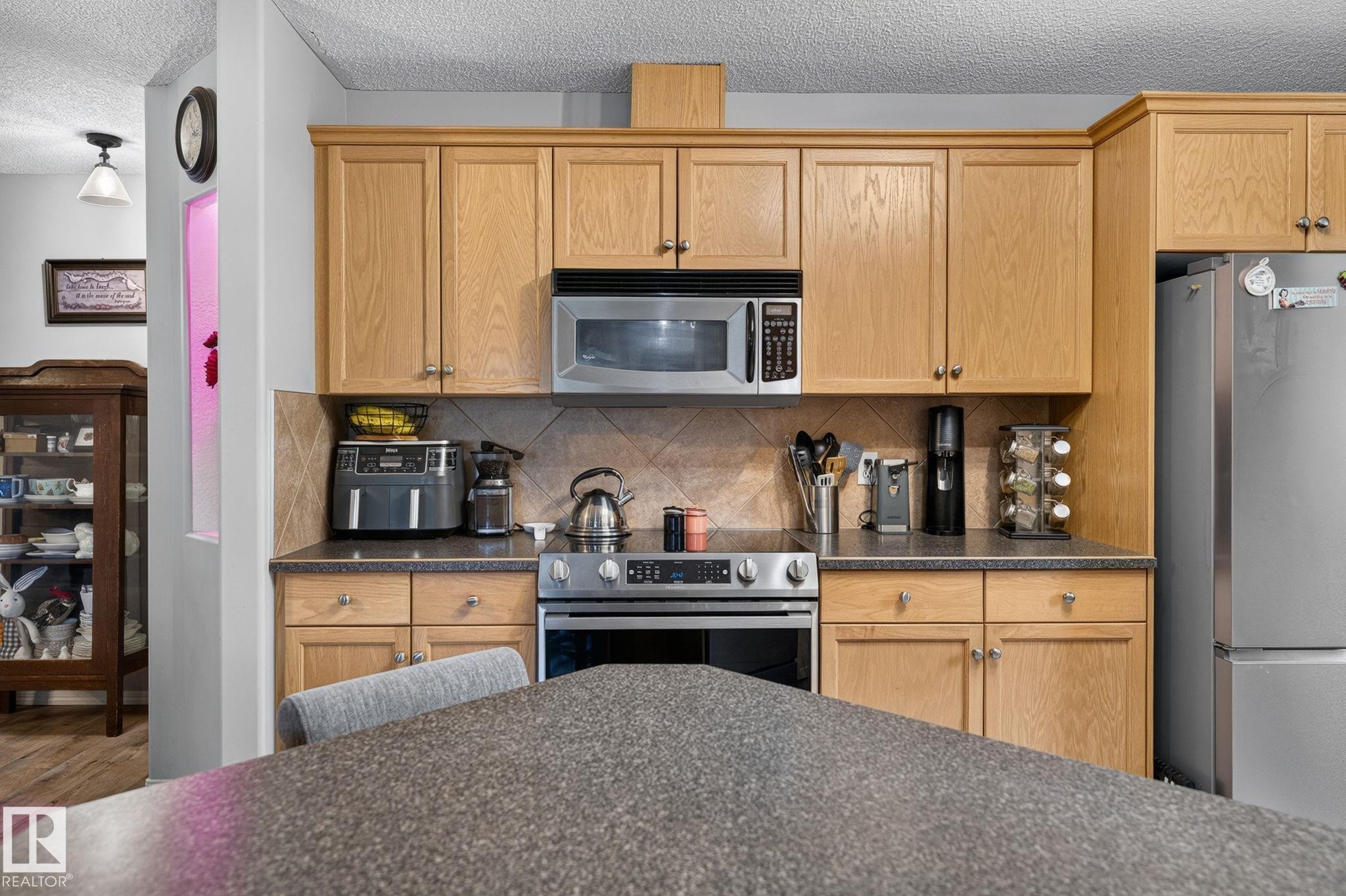 Kitchen featuring wood-finish cabinetry, stainless steel appliances, a textured tile backsplash, and a dark speckled countertop - 122 279 Suder Greens Drive, Edmonton, AB