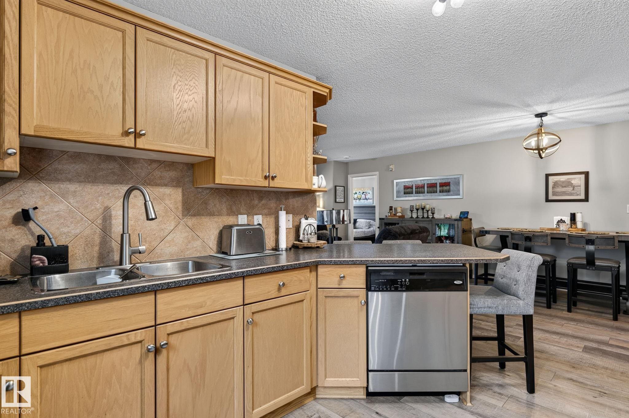 Kitchen featuring light wood cabinetry with silver hardware, dual basin sink with gooseneck faucet, tiled backsplash, and stone-finish countertops - 122 279 Suder Greens Drive, Edmonton, AB