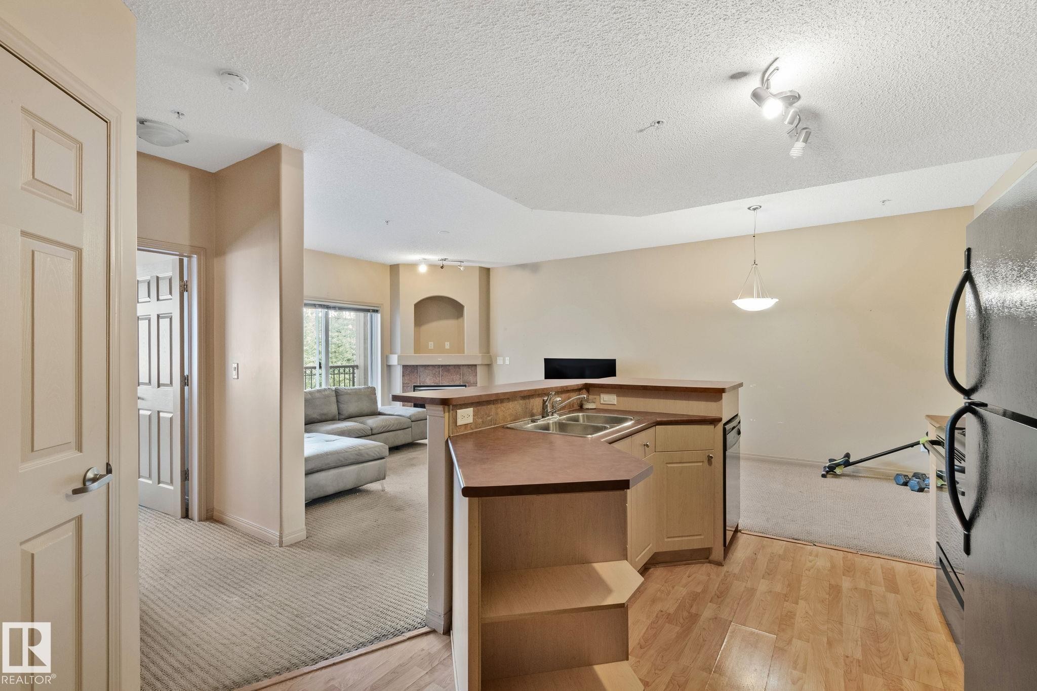 Open-concept living area featuring a kitchen island with dual sinks, wood-finish flooring, and a fireplace with a stone surround - 209 5280 Terwillegar Boulevard, Edmonton, AB - Indoor Photo Showing Other Room