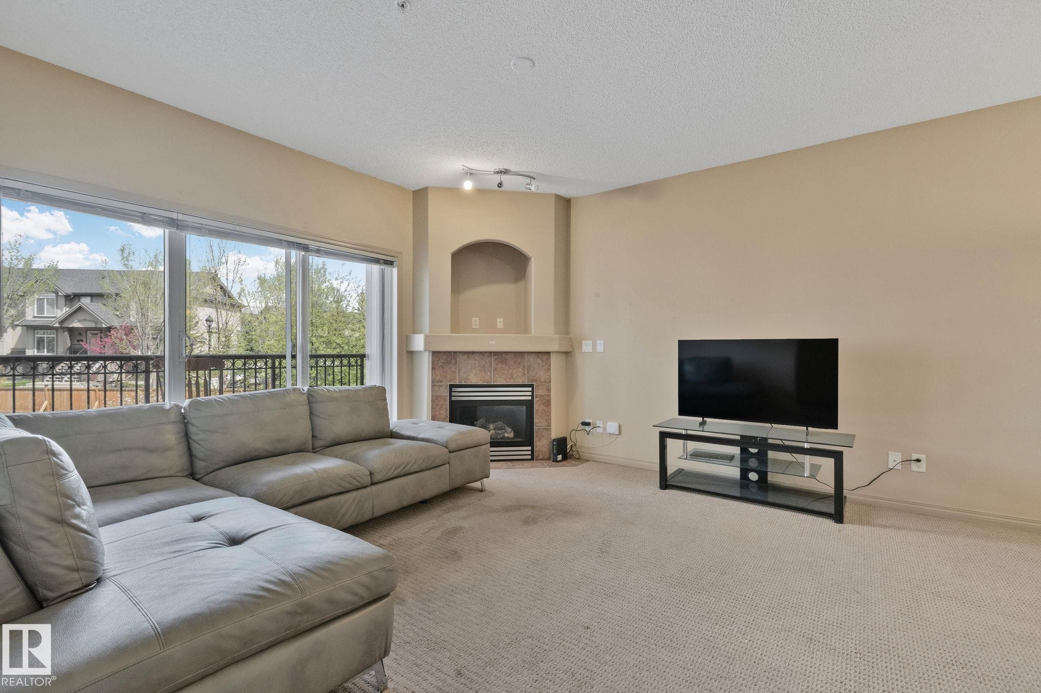 Living area with corner gas fireplace featuring a stone surround and mantel, sliding glass doors opening to a balcony, and neutral carpeting - 209 5280 Terwillegar Boulevard, Edmonton, AB - Indoor Photo Showing Living Room With Fireplace