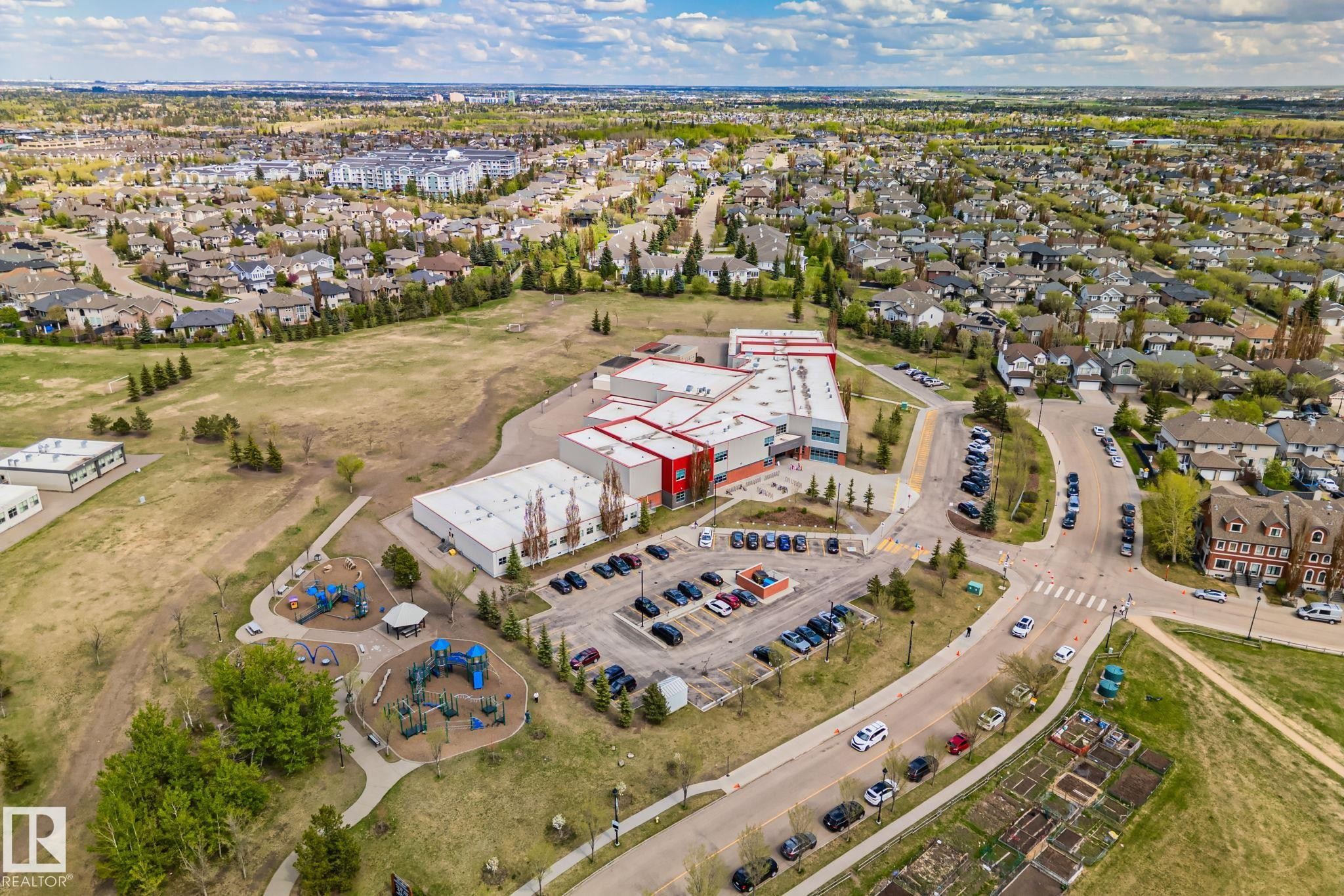 Expansive aerial perspective showcasing a residential development with numerous single-family homes and multi-story buildings, featuring a central school building with parking, multiple playgrounds, and open green spaces - 209 5280 Terwillegar Boulevard, Edmonton, AB - Outdoor With View