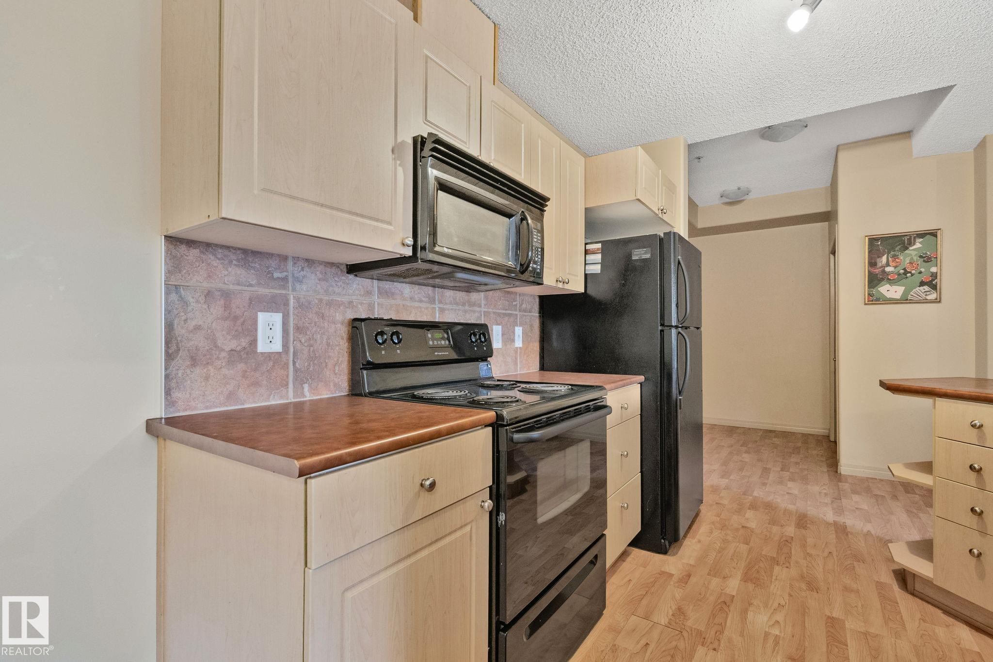 Kitchen featuring light wood-finish cabinetry, black appliances including a built-in microwave and range, and warm-toned countertops - 209 5280 Terwillegar Boulevard, Edmonton, AB - Indoor Photo Showing Kitchen