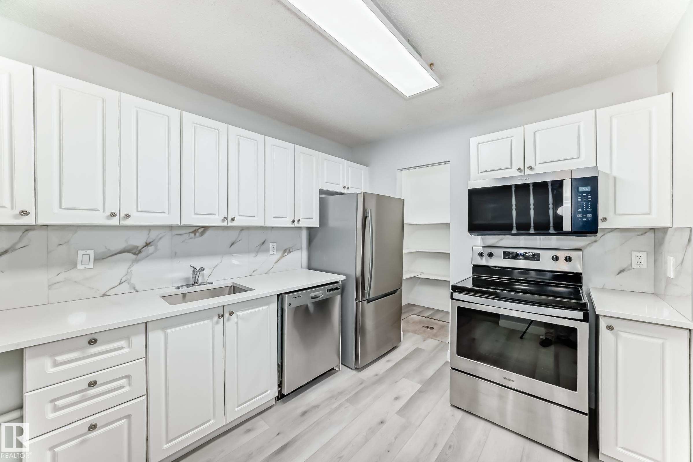 Kitchen featuring bright white cabinetry, stainless steel appliances, marble-patterned backsplash, light wood-finish flooring, and ample counter space - 23 1503 Mill Woods Road E, Edmonton, AB