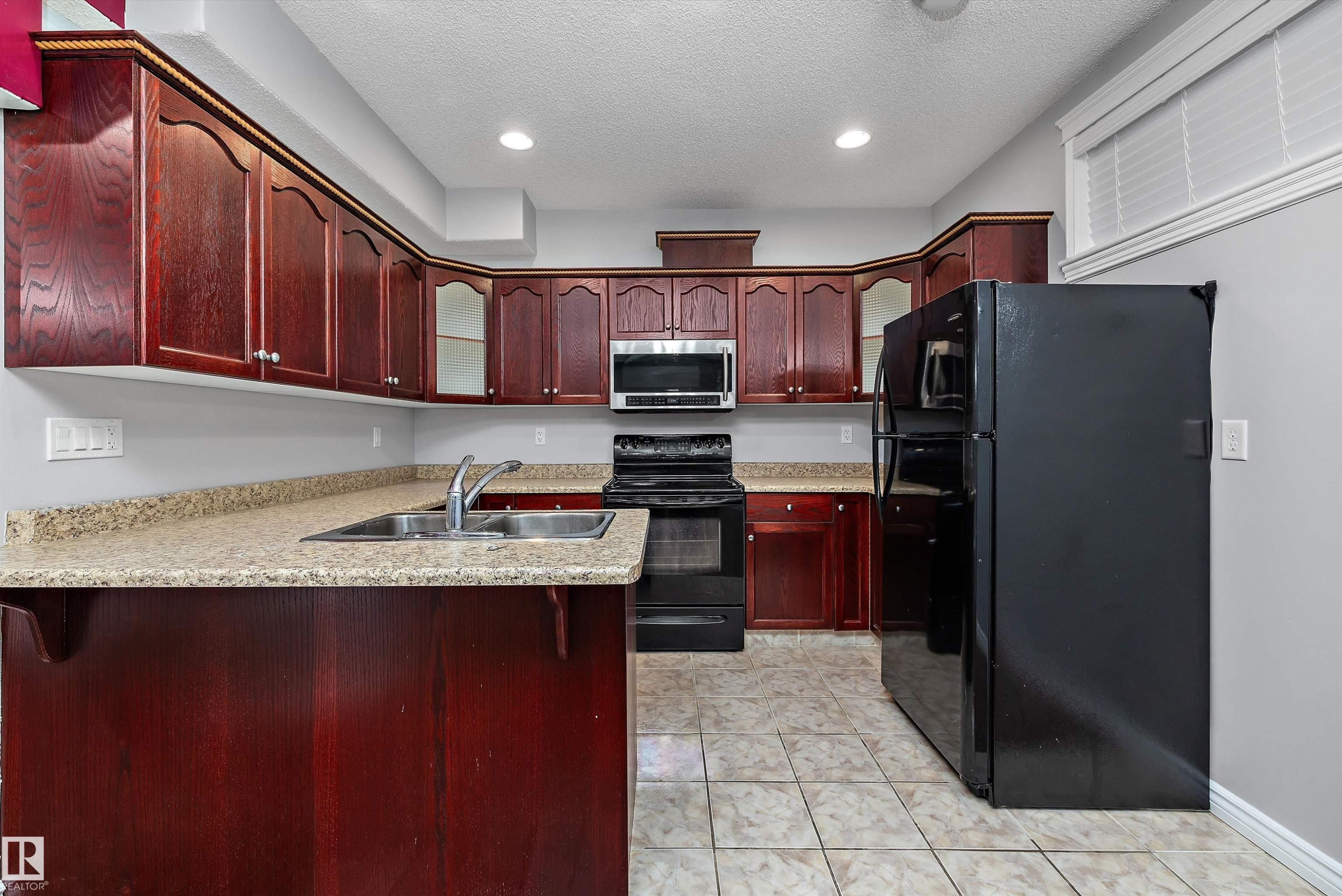 Kitchen featuring dark wood cabinetry, light-toned countertops, a double basin stainless steel sink, and a full suite of black appliances, including a microwave and refrigerator - 4831 154 Avenue, Edmonton, AB