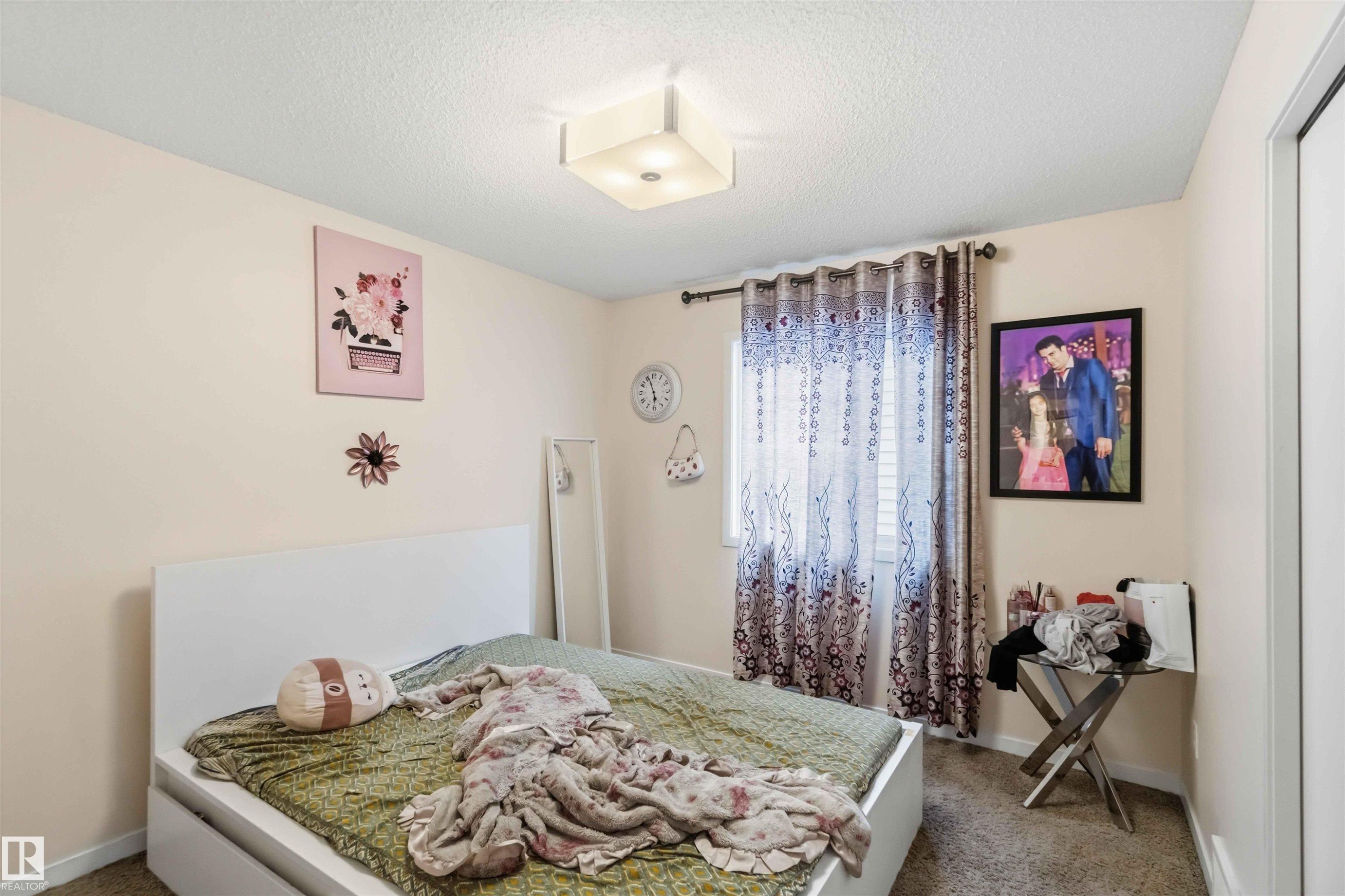 Bedroom featuring a white platform bed frame, a single window with blinds and patterned drapes, textured carpeting, and a flush mount ceiling light fixture - 5120 19 Avenue, Edmonton, AB
