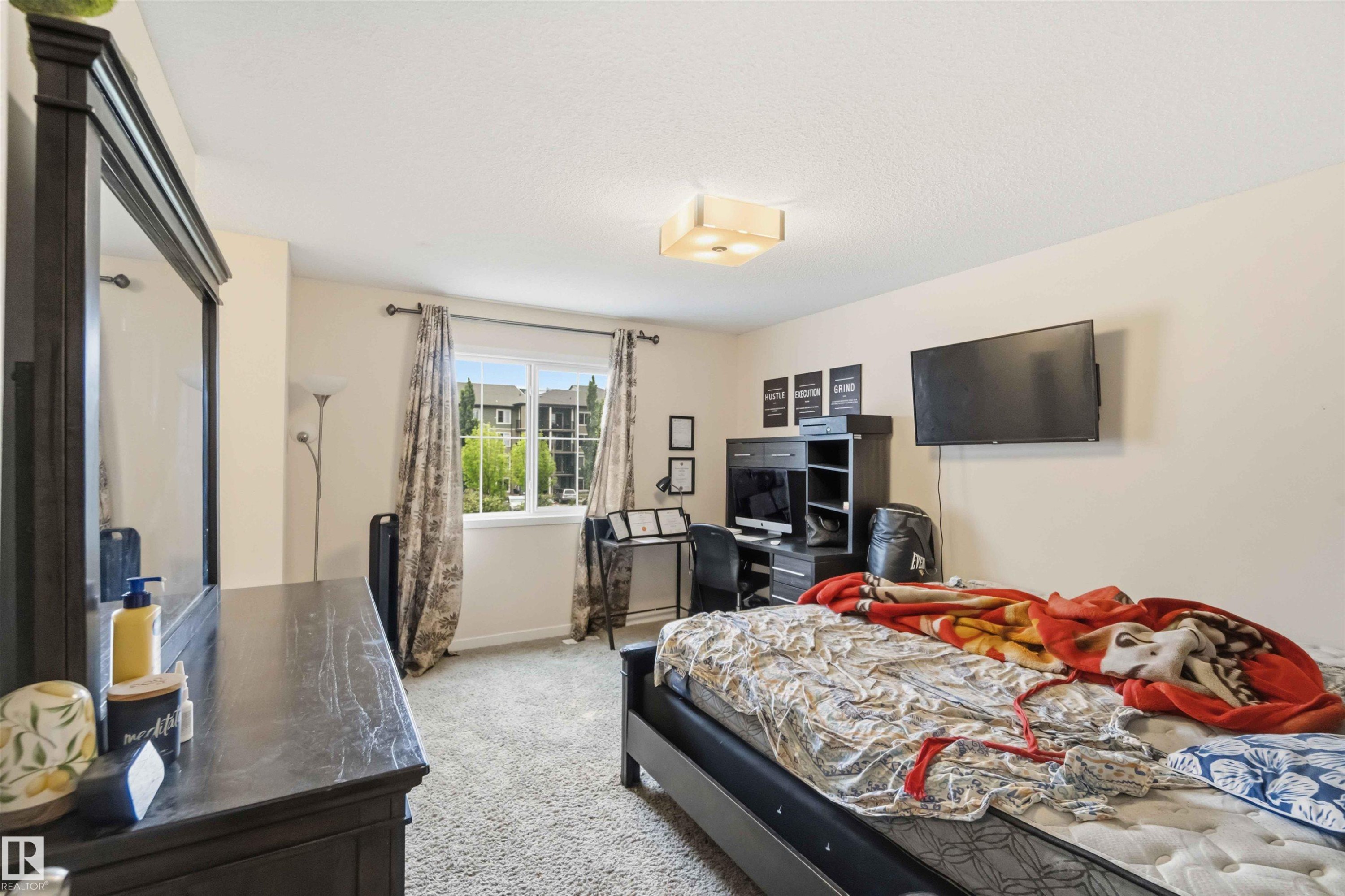 Carpeted room with a ceiling-mounted light fixture, a window with exterior views, and a wall-mounted television - 5120 19 Avenue, Edmonton, AB