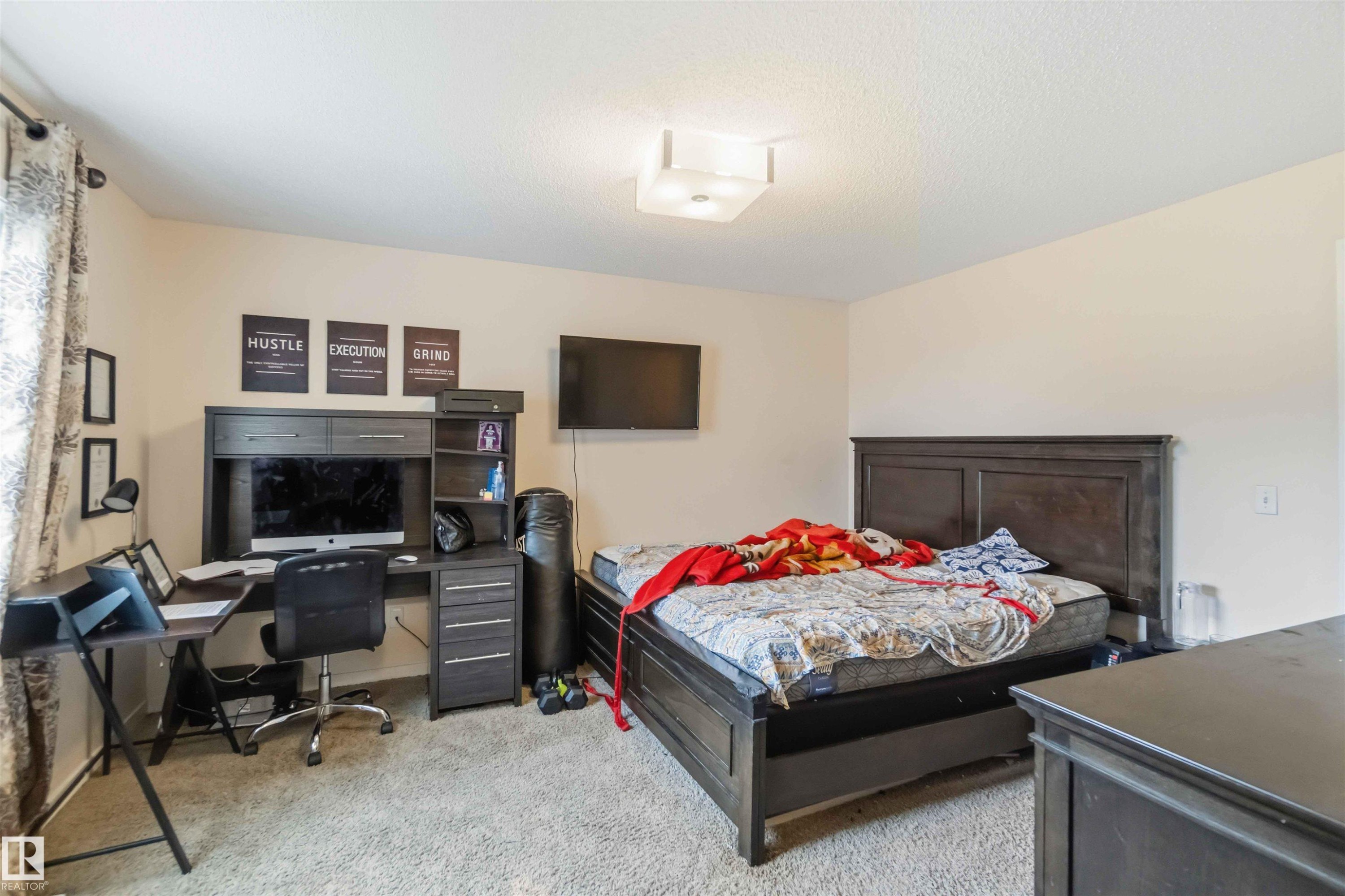 Carpeted bedroom featuring neutral-toned walls, a built-in flush mount ceiling light fixture, and a window with patterned drapes - 5120 19 Avenue, Edmonton, AB