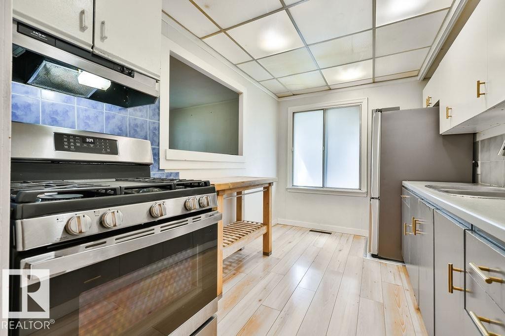 Stainless steel gas range with overhead exhaust hood, blue ceramic tile backsplash, and light wood-finish flooring - 8318 80 Avenue, Edmonton, AB